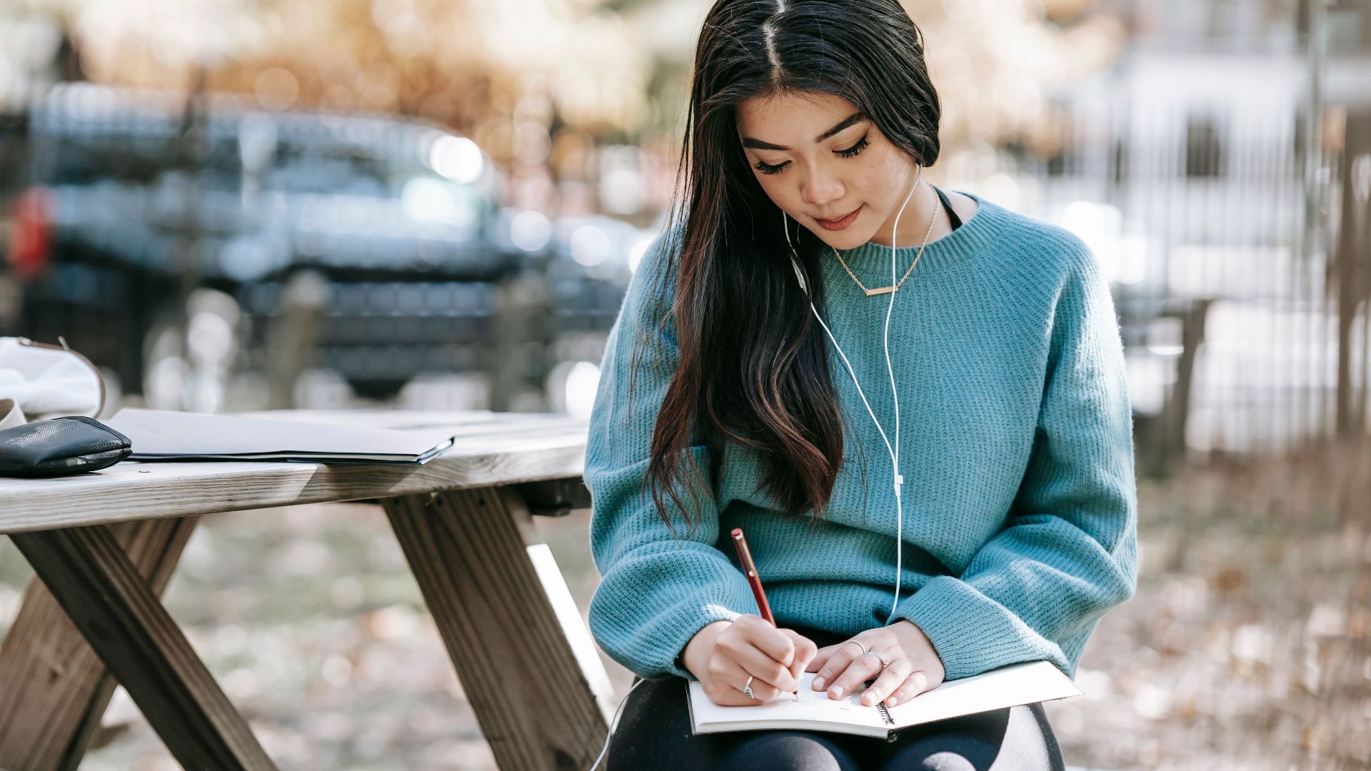 Young concentrated ethnic female taking notes in copybook while listening to music in earphones at table on blurred background of street
