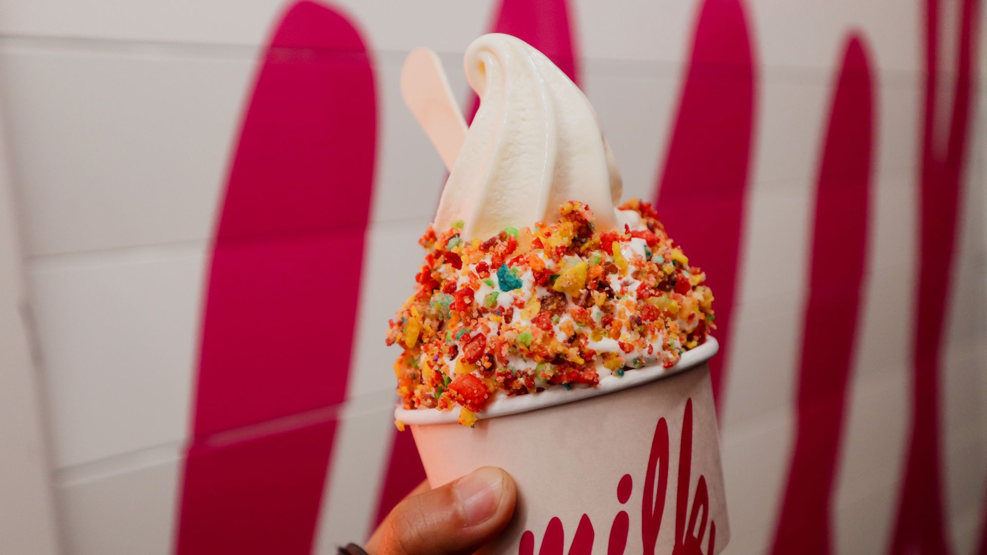 Hand holding a vanilla soft serve ice cream with colorful toppings in a vibrant setting.