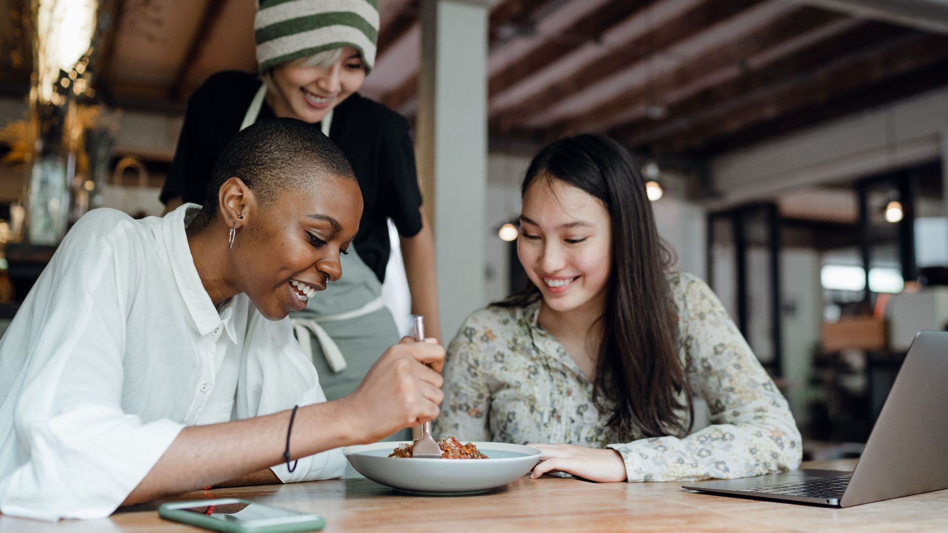 Positive joyful diverse women in casual clothes tasting new menu dish while sitting at table with laptop near smiling waitress