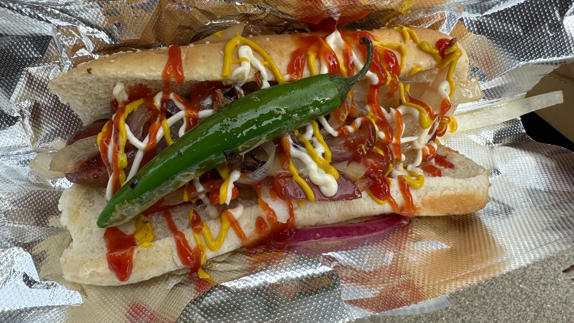 A bacon-wrapped hot dog with onions, peppers, mayo, ketchup and mustard.