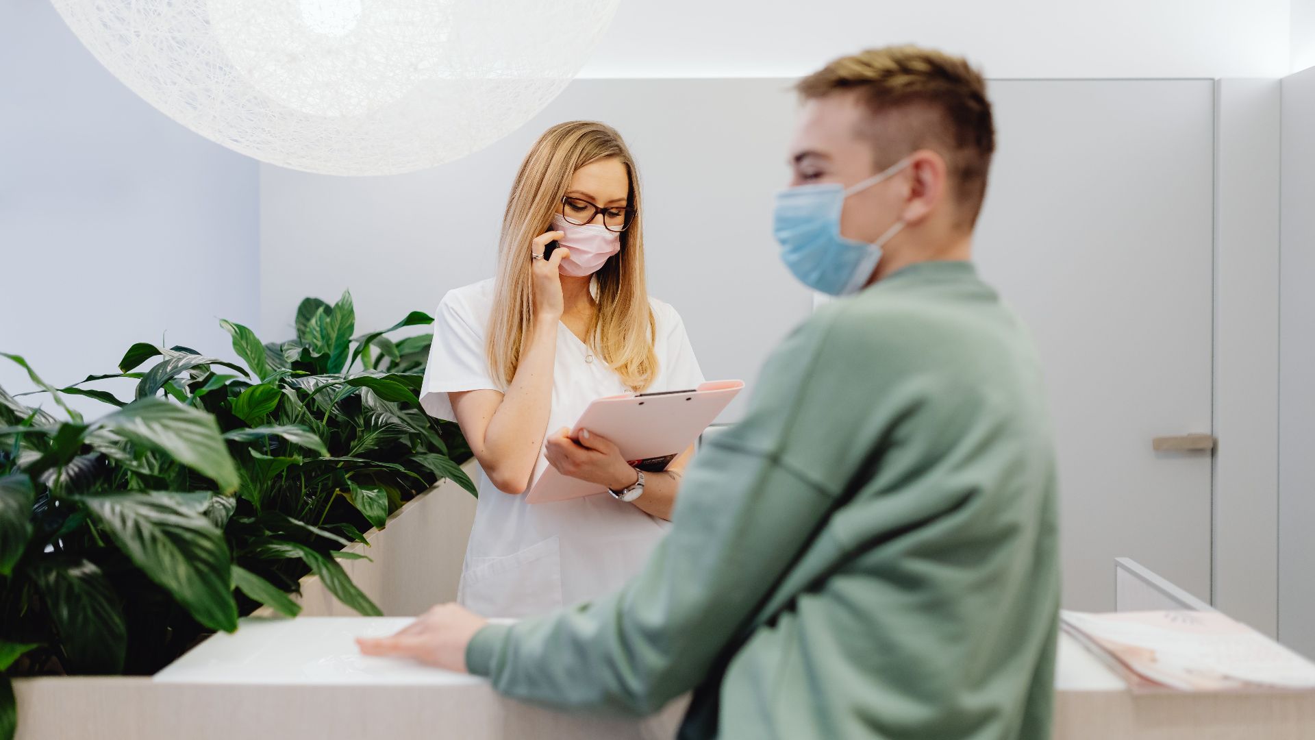 Healthcare worker and patient wearing masks at clinic front desk during pandemic, highlighting safety measures.