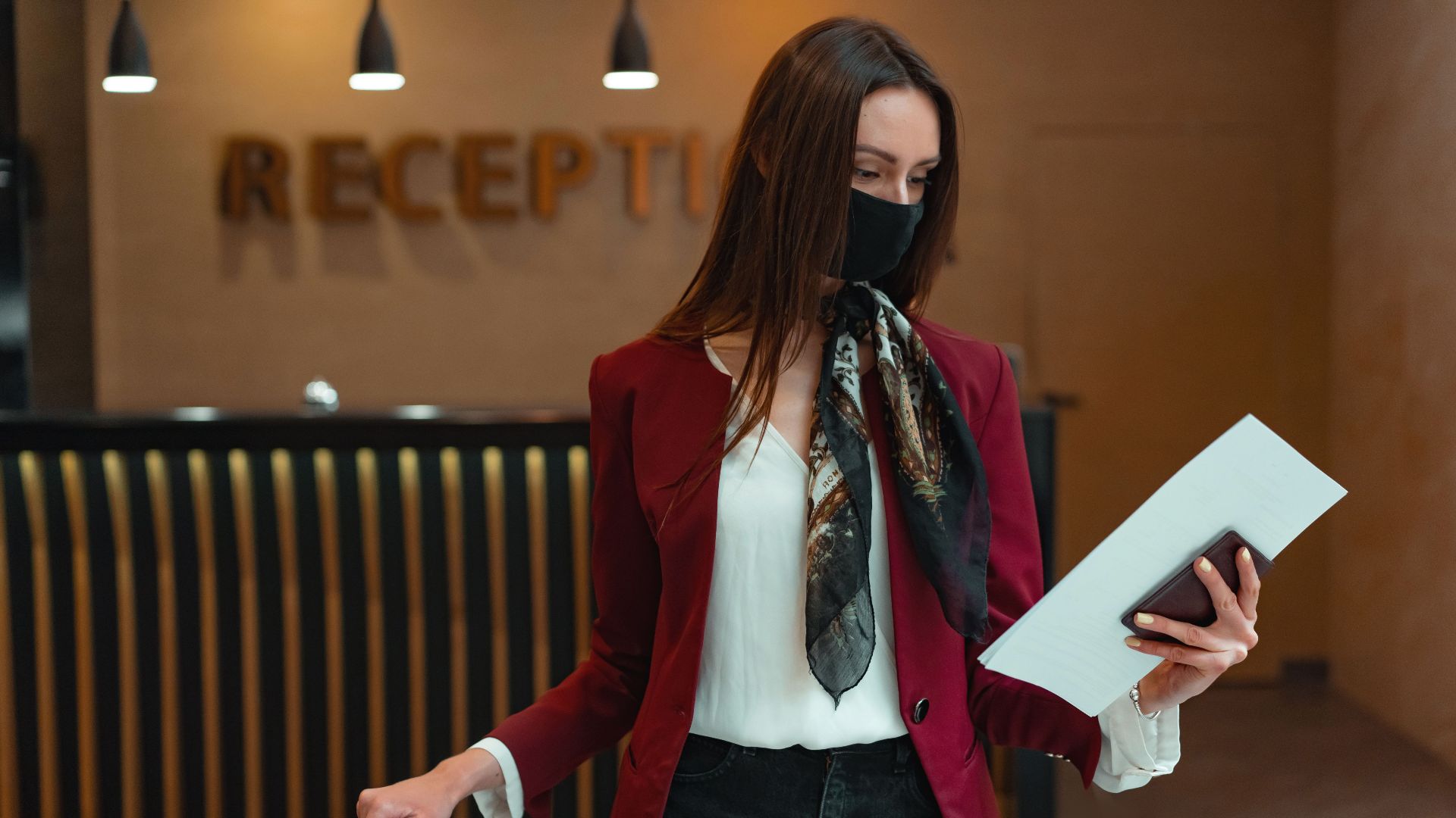 Woman in red blazer at hotel reception, holding luggage and documents, wearing a face mask.