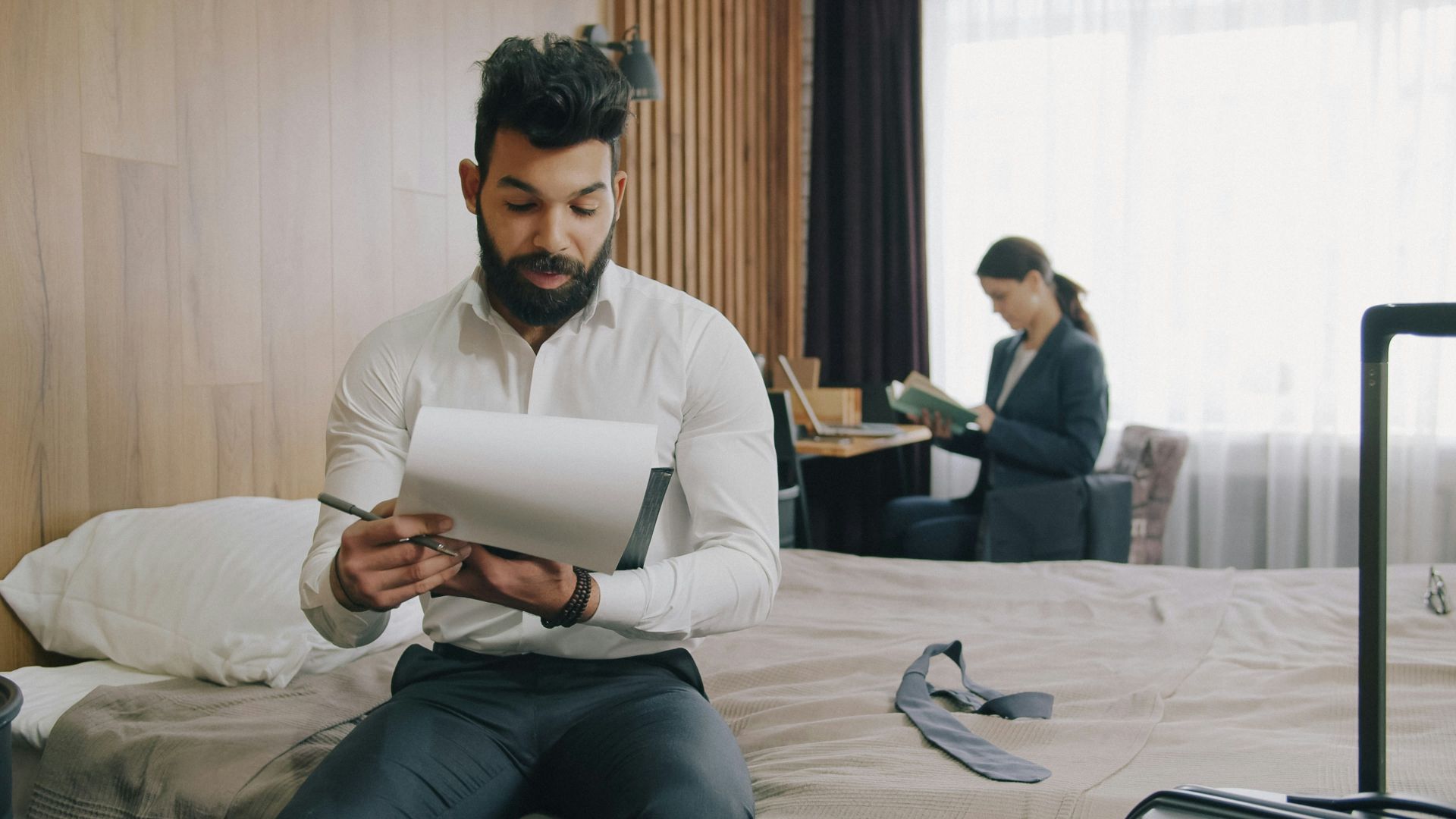 A man in a white shirt writing in a notebook on a hotel bed