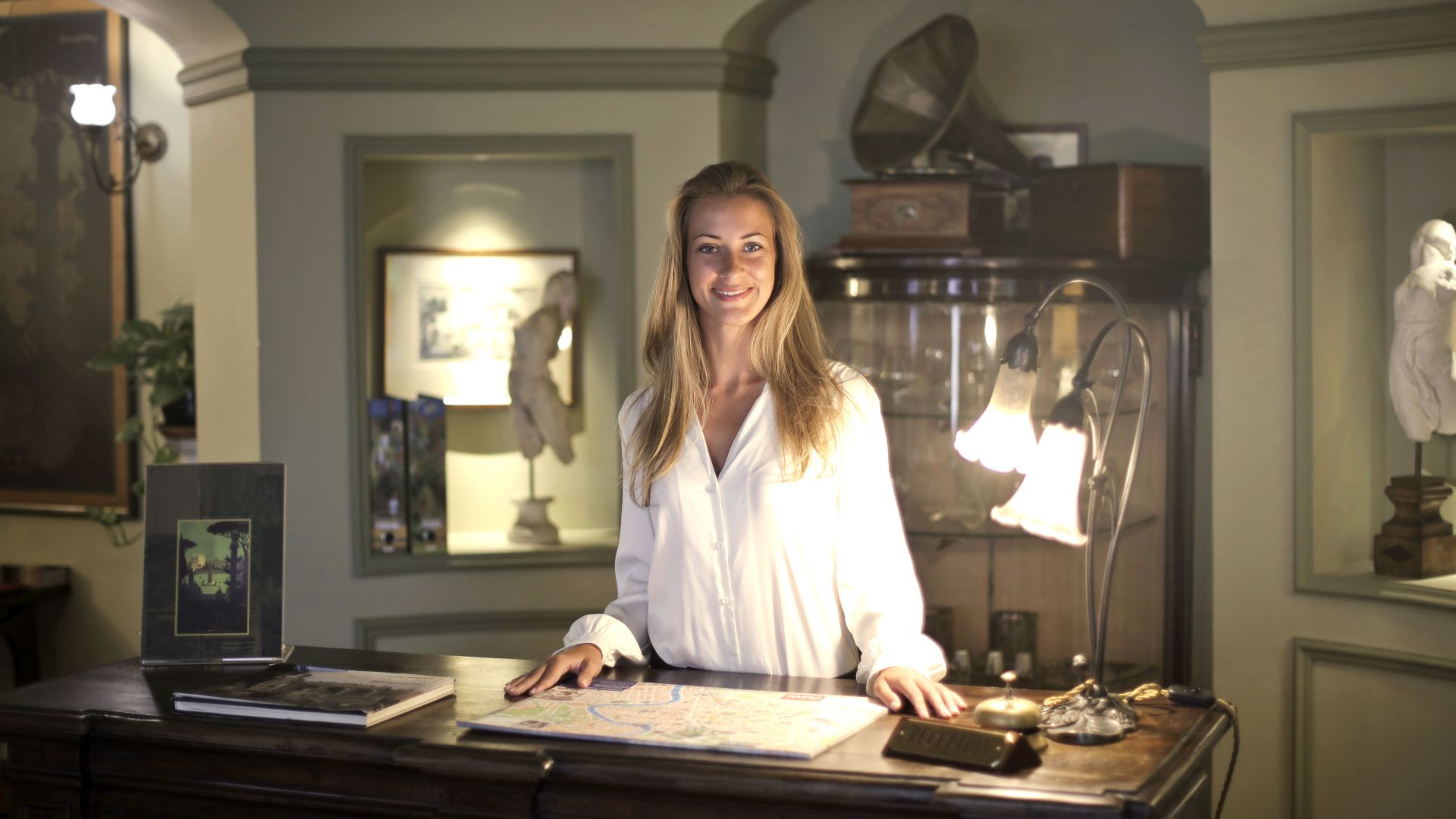 Smiling female receptionist wearing elegant clothes standing at reception desk in old fashioned interior of hotel and looking at camera