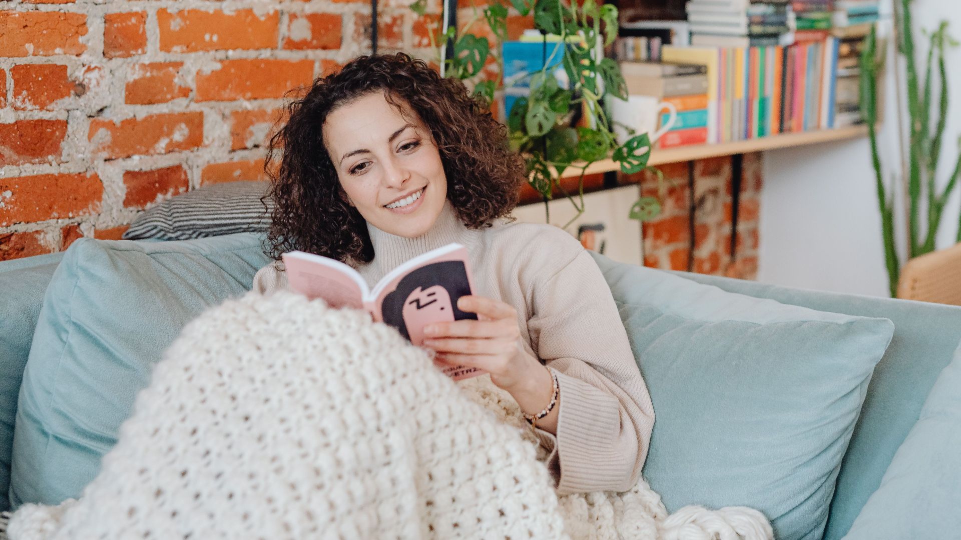 A smiling woman reads on a sofa in a cozy room with bookshelves and plants.
