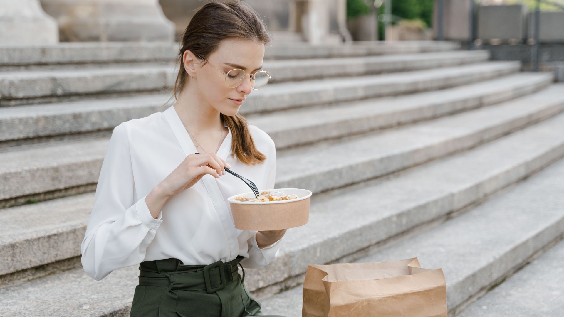Woman seated outdoors on steps, enjoying a takeout meal from a paper bowl, wearing business casual attire.