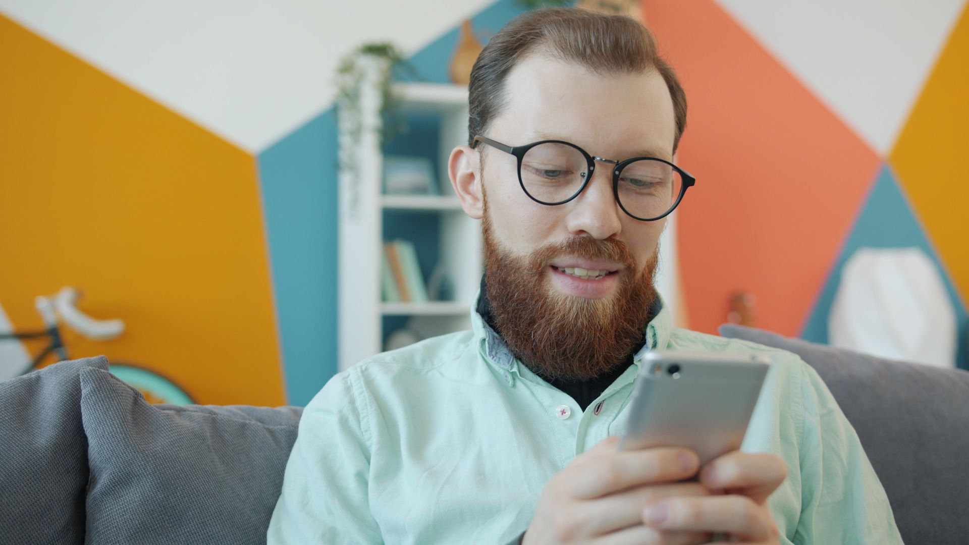 Man with glasses and beard using smartphone indoors with vibrant decor.