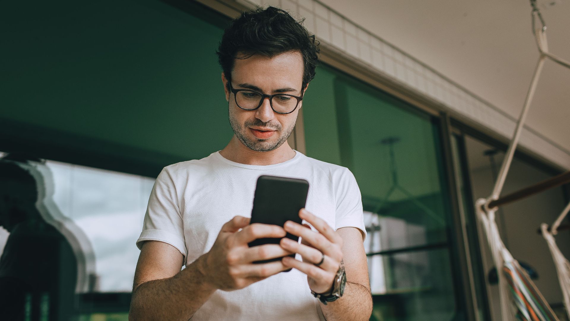 Young man standing on a balcony, using a smartphone, reflecting in the window.