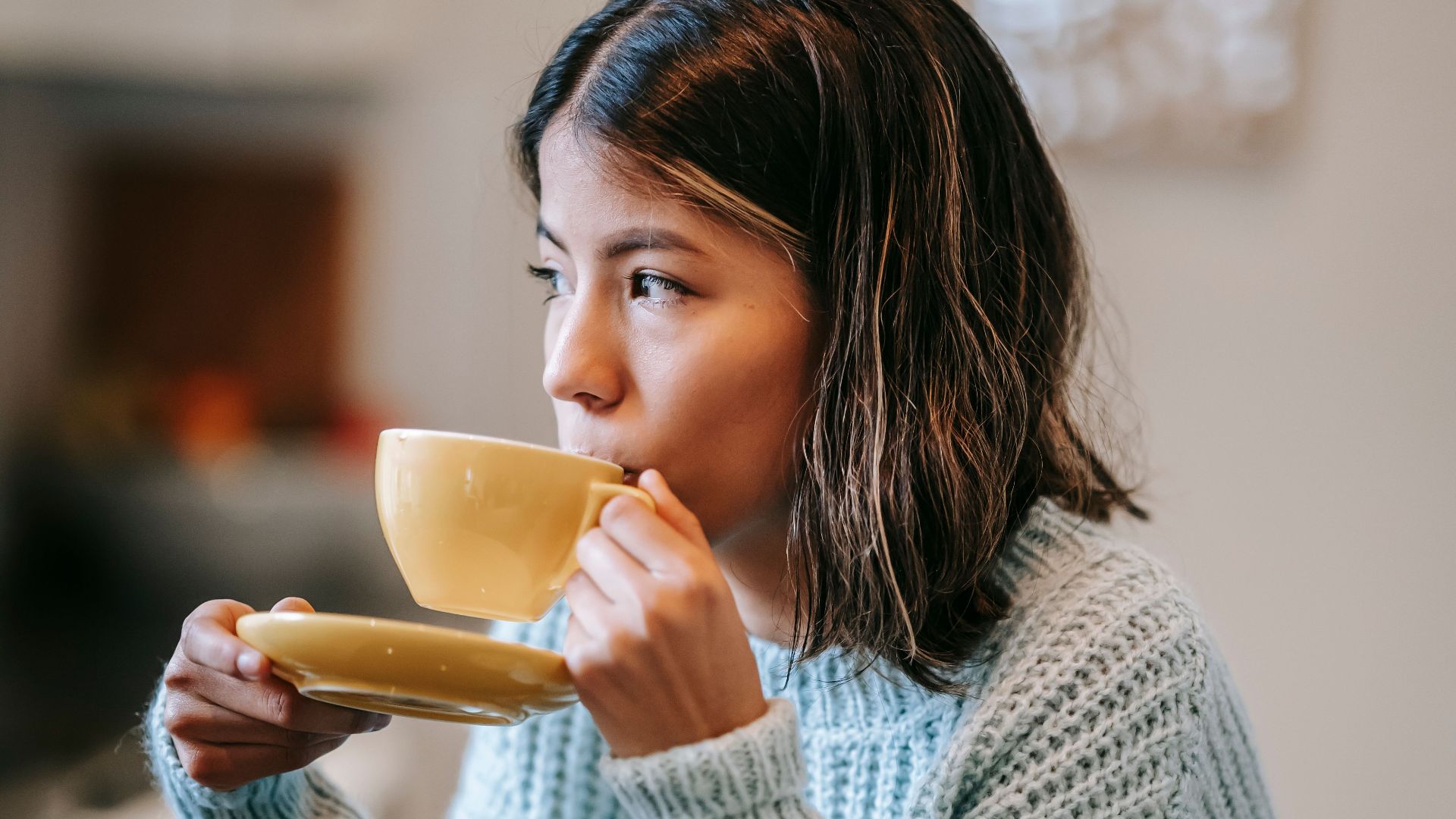 Young female with short hair drinking cup of aromatic hot coffee and looking away at table with sweet delicious croissant