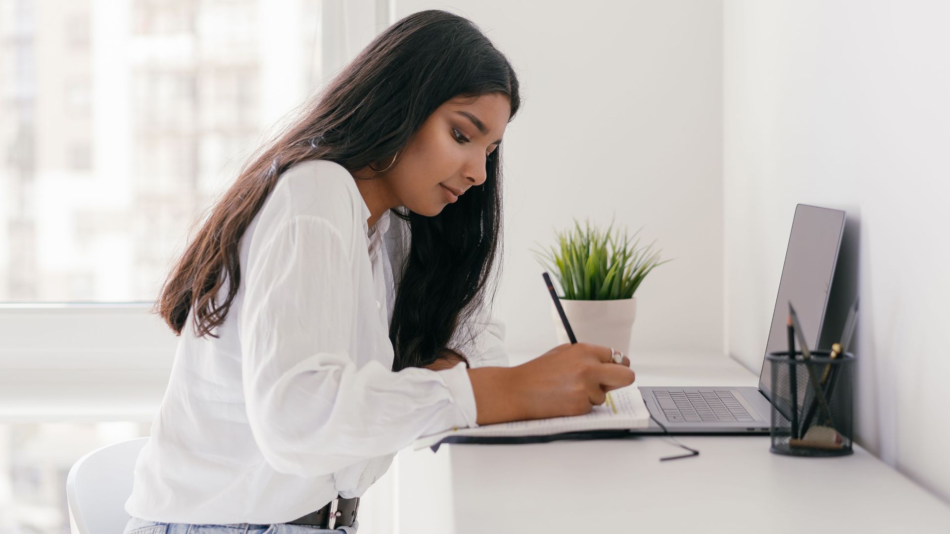 Young woman studying at a bright home desk with a laptop and notebook, focused on writing.