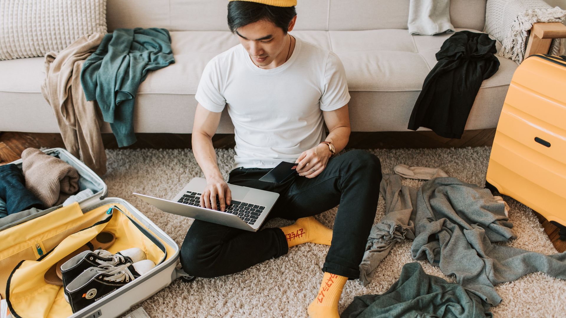 Man multitasking by packing luggage while working on a laptop in living room.