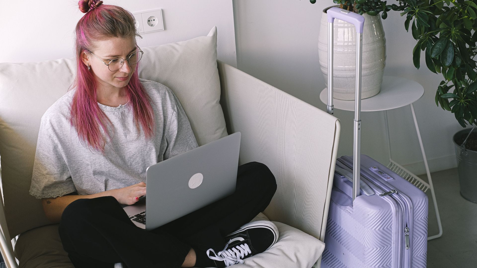 Casual young woman with pink hair works on laptop in comfortable indoor setting, travel suitcase nearby.