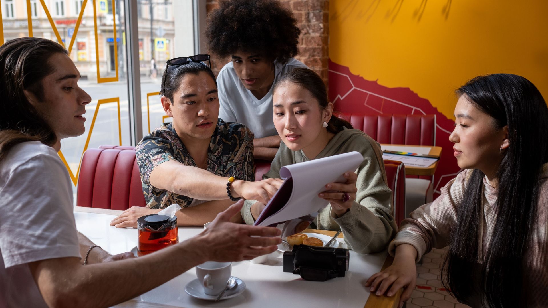 A multicultural group of young adults engaged in conversation around a table in a cafe, focused on a document.