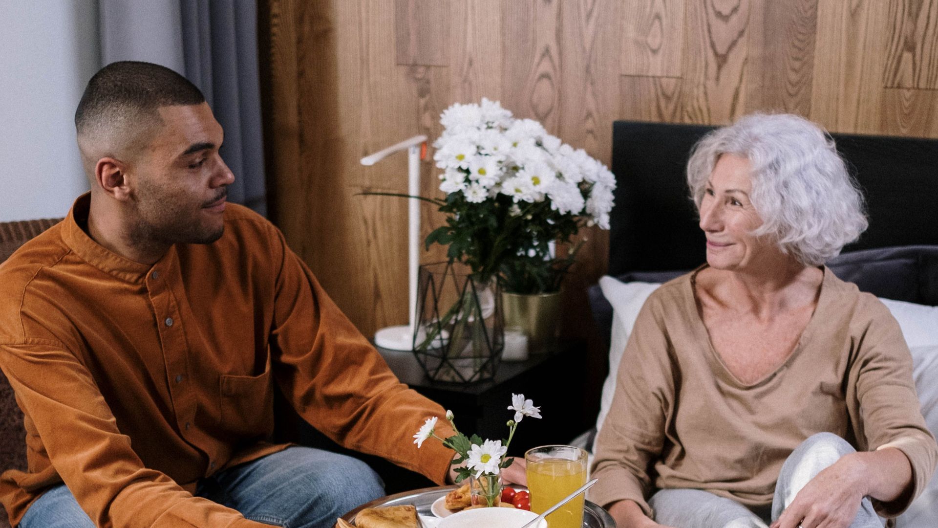 A young man serving breakfast in bed to his elderly grandmother with a warm smile.