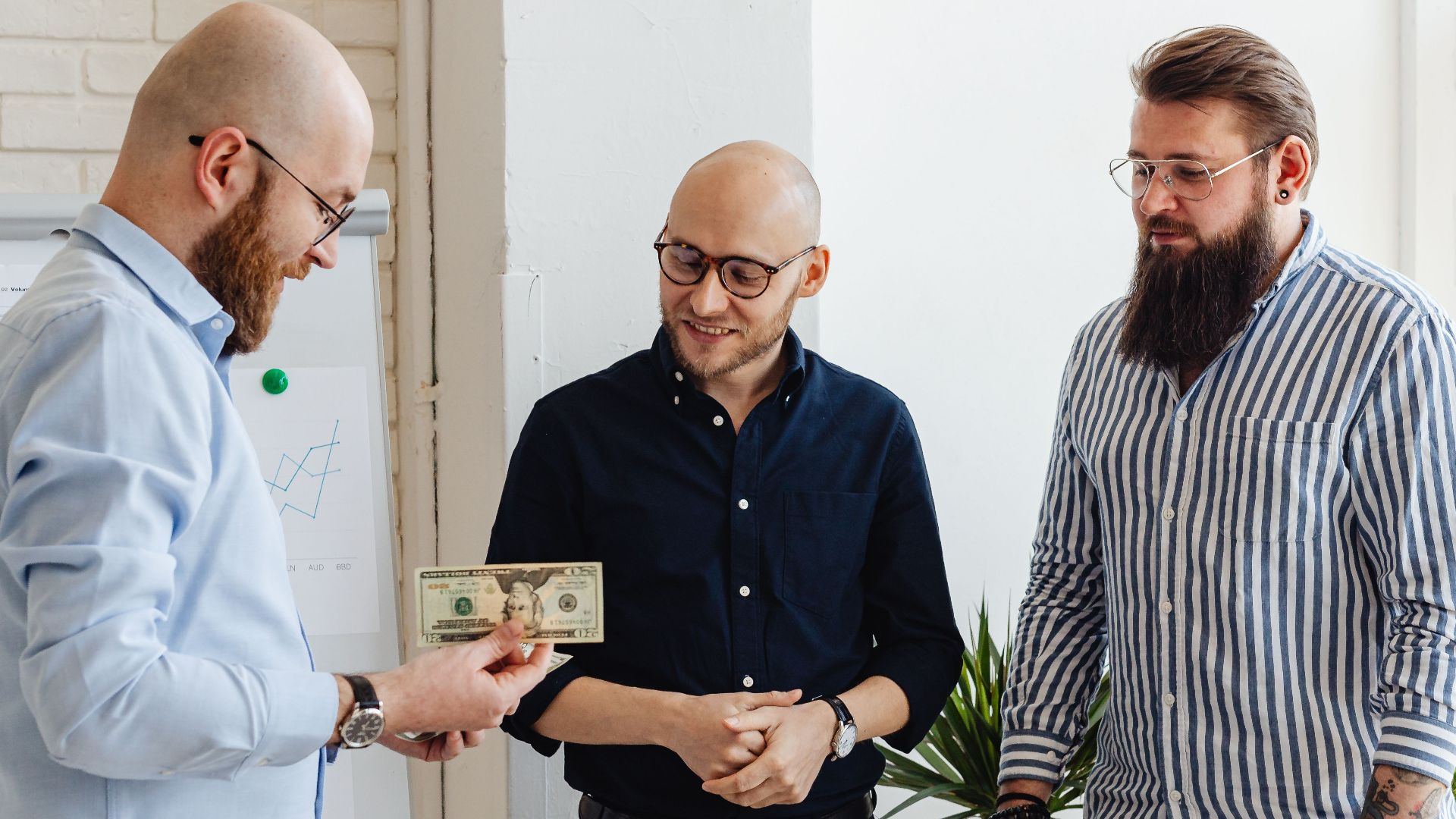 Three businessmen exchanging money in a modern office with financial charts.
