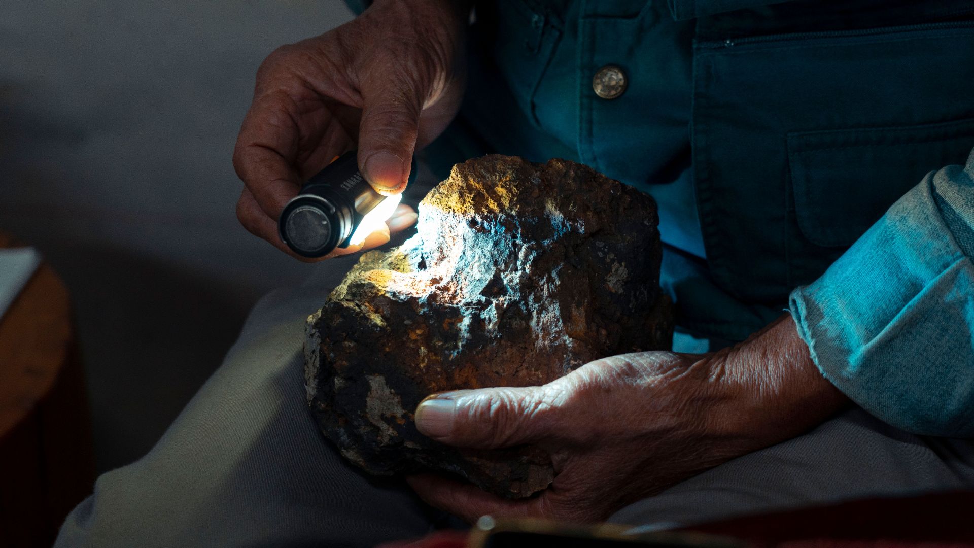 Detailed close-up of hands using a flashlight to examine a geological stone indoors.