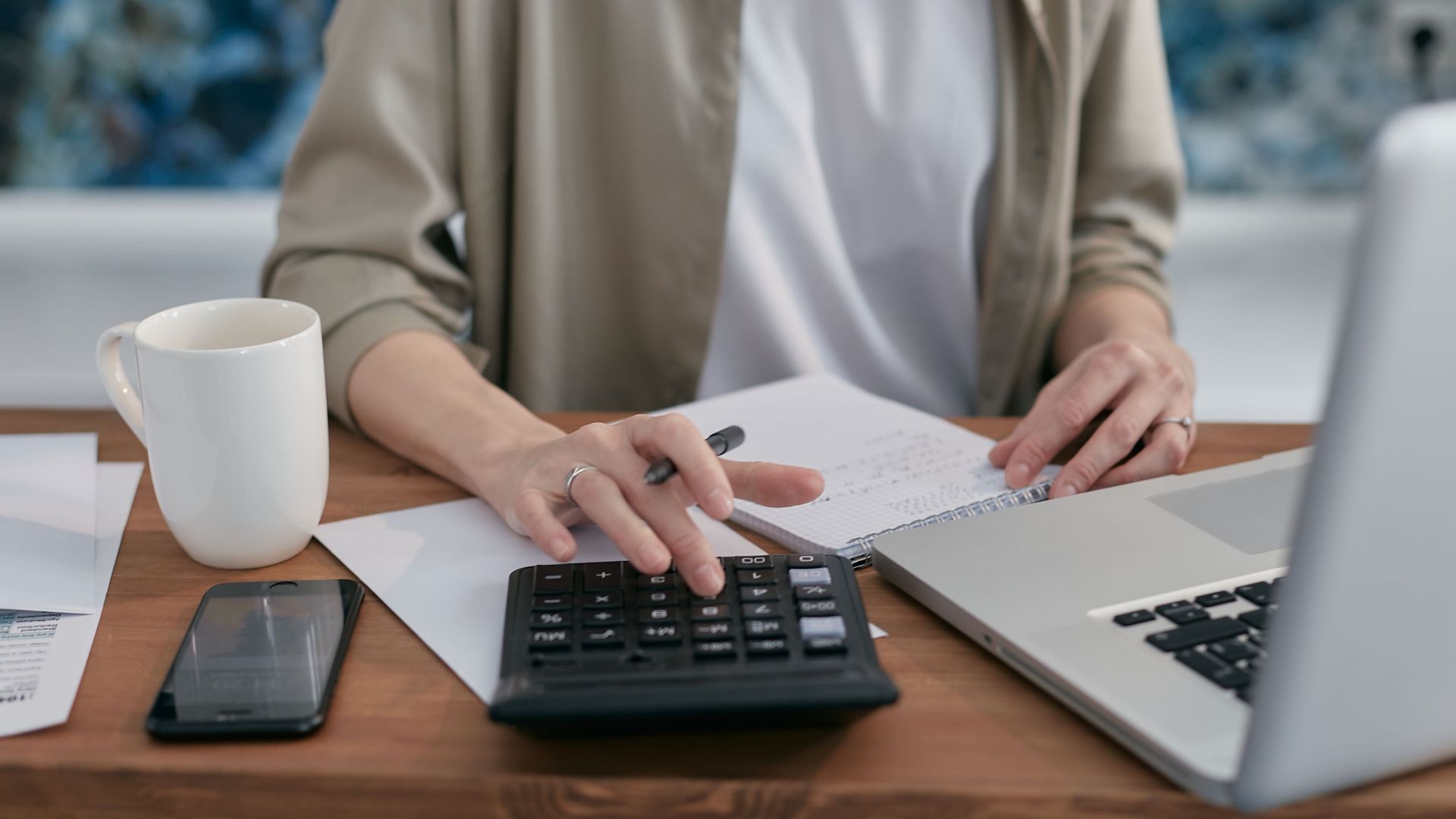A person using a calculator and laptop while taking notes at a wooden desk.