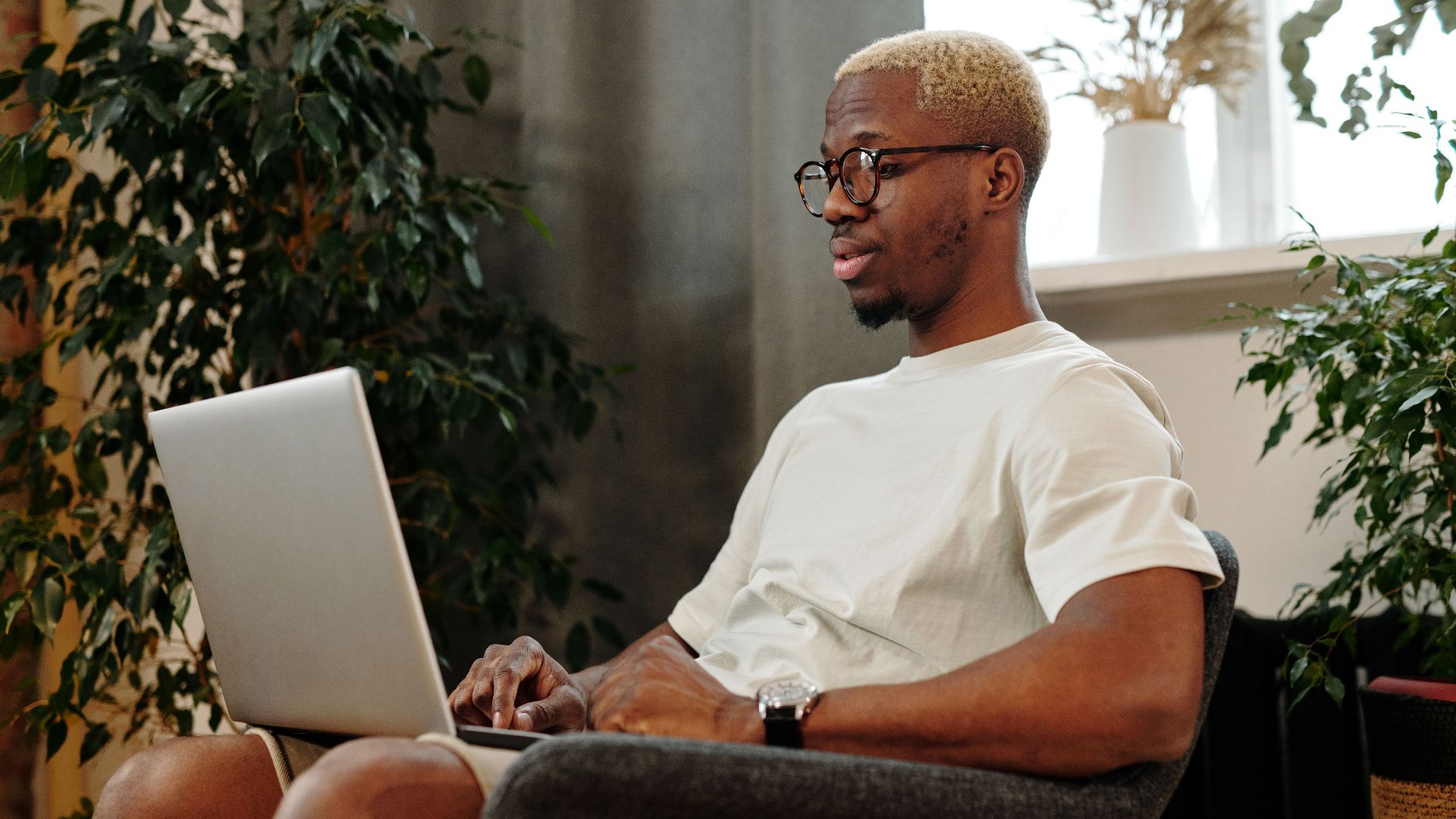 Focused young man with glasses working on a laptop in a cozy indoor space surrounded by plants.