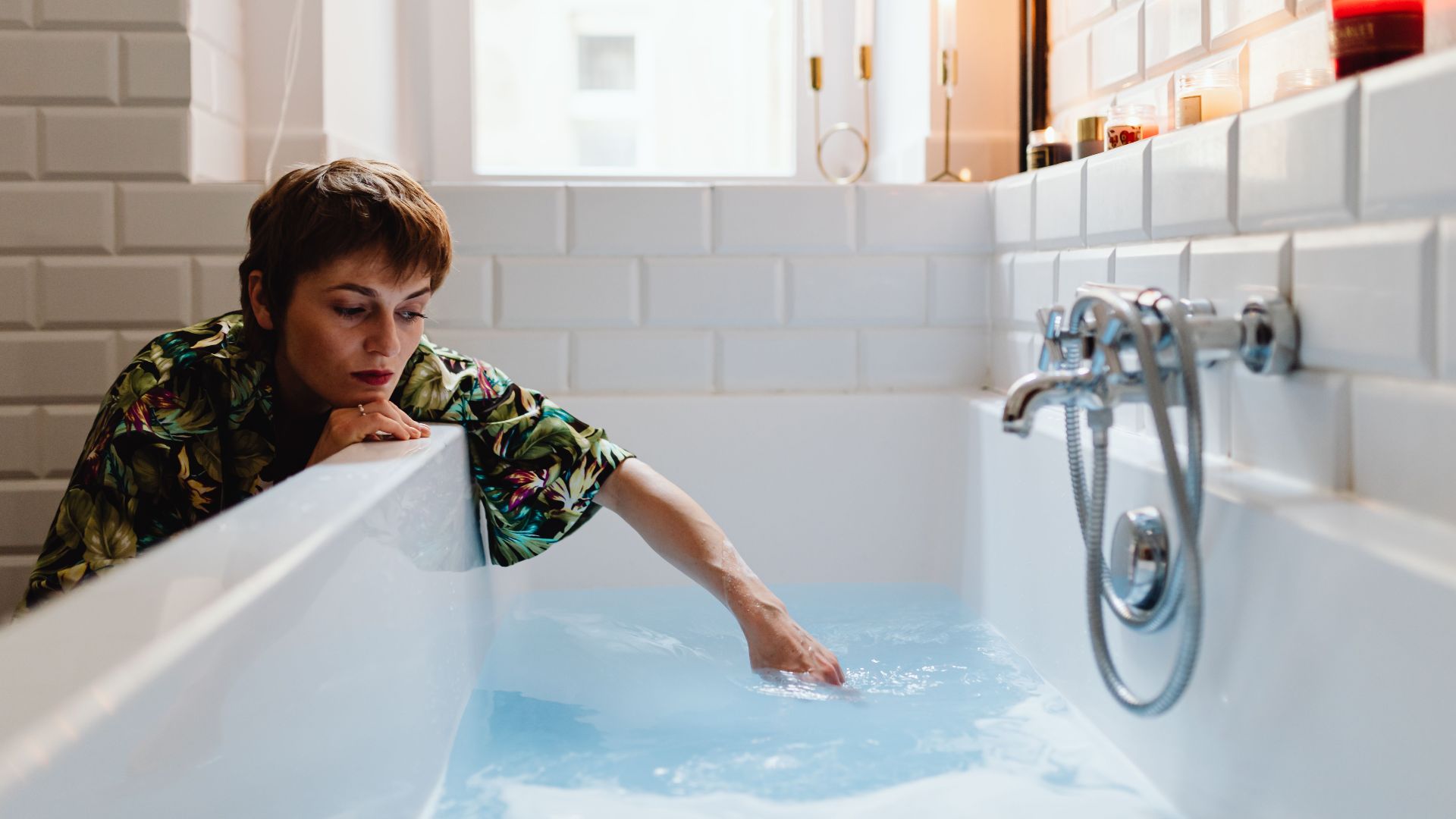 A woman in a serene bathroom touches the water in a modern white tiled bathtub.