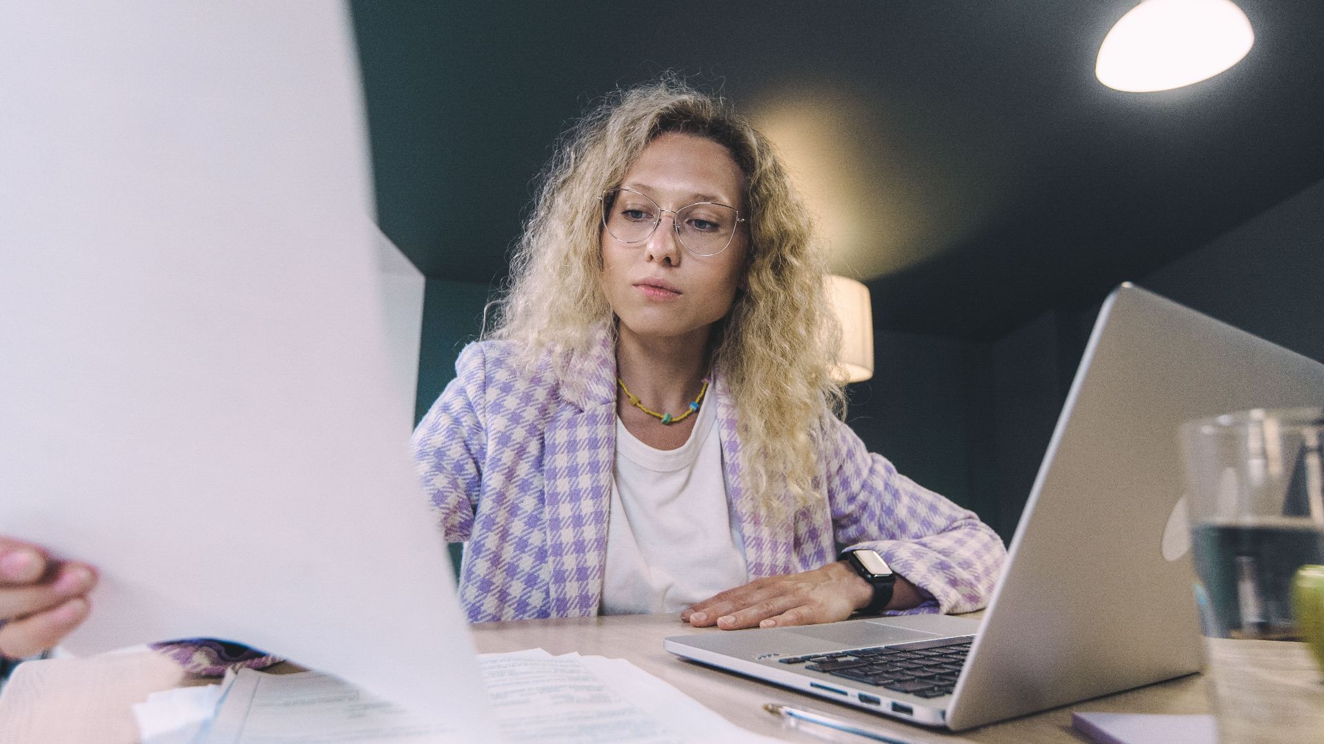 Woman in an office environment reviewing documents with focus, surrounded by technology.
