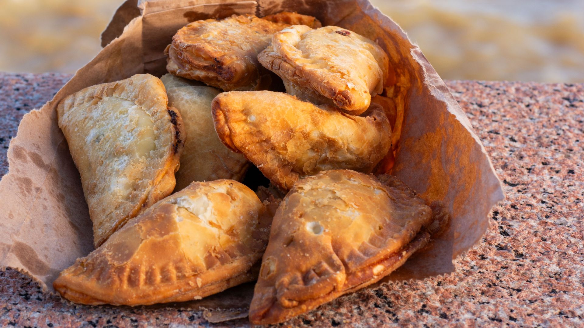 Brown paper bag filled with golden, crispy empanadas sitting on a stone surface by the Montevideo seaside.