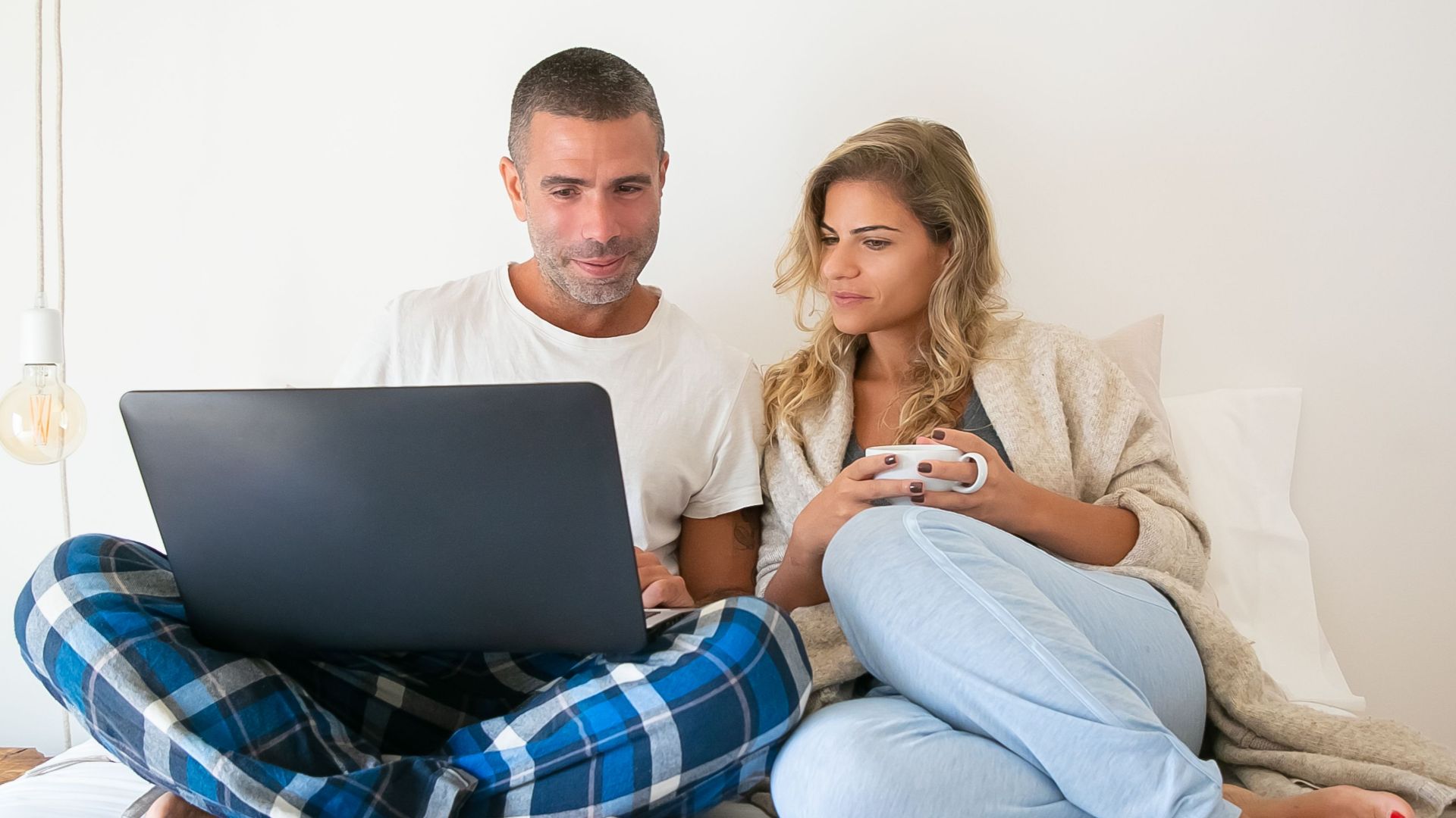 A couple in casual wear working remotely from bed, sharing a laptop in a cozy home setting.