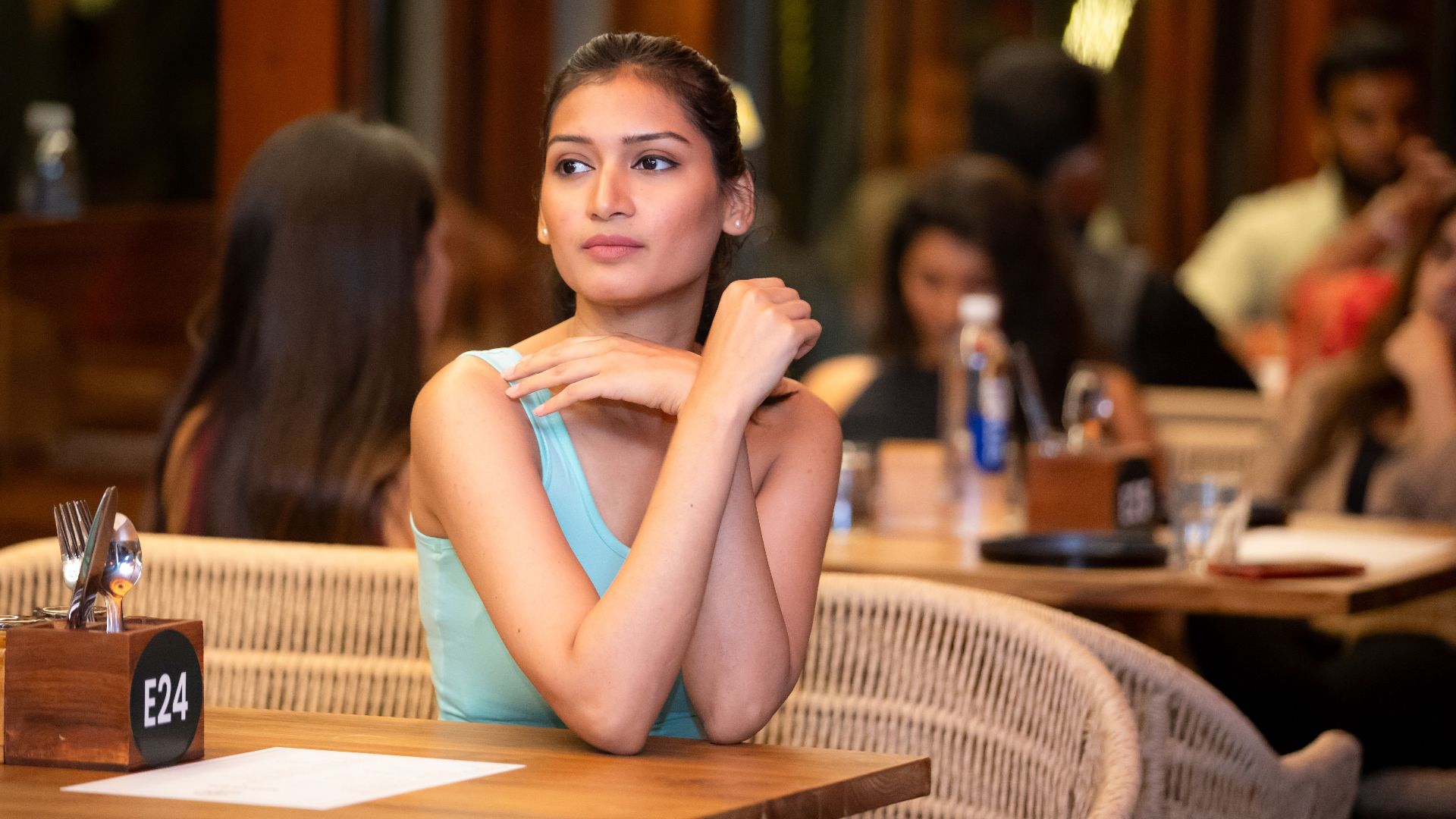 A young woman in a turquoise top sits in a lively, crowded restaurant, appearing thoughtful.
