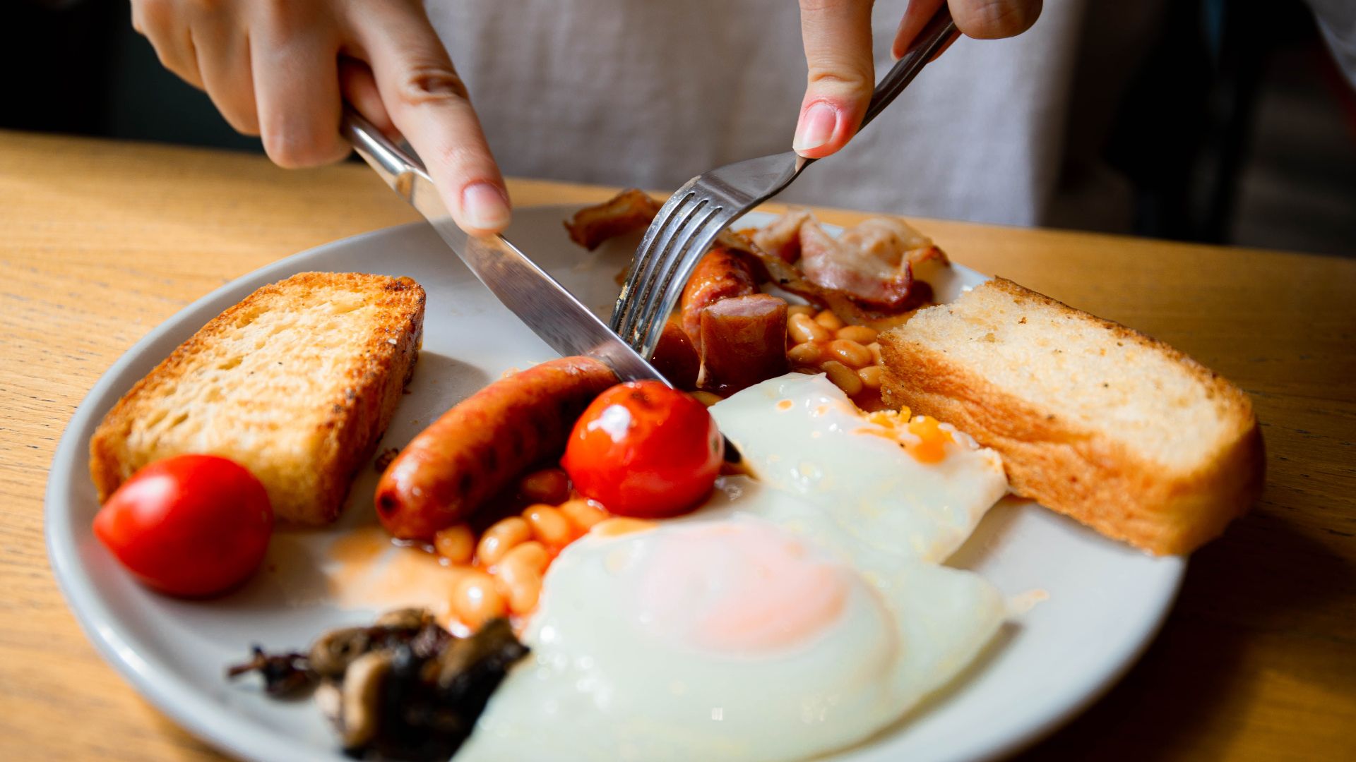Hand slicing a full English breakfast with bacon, sausage, eggs, and tomatoes.