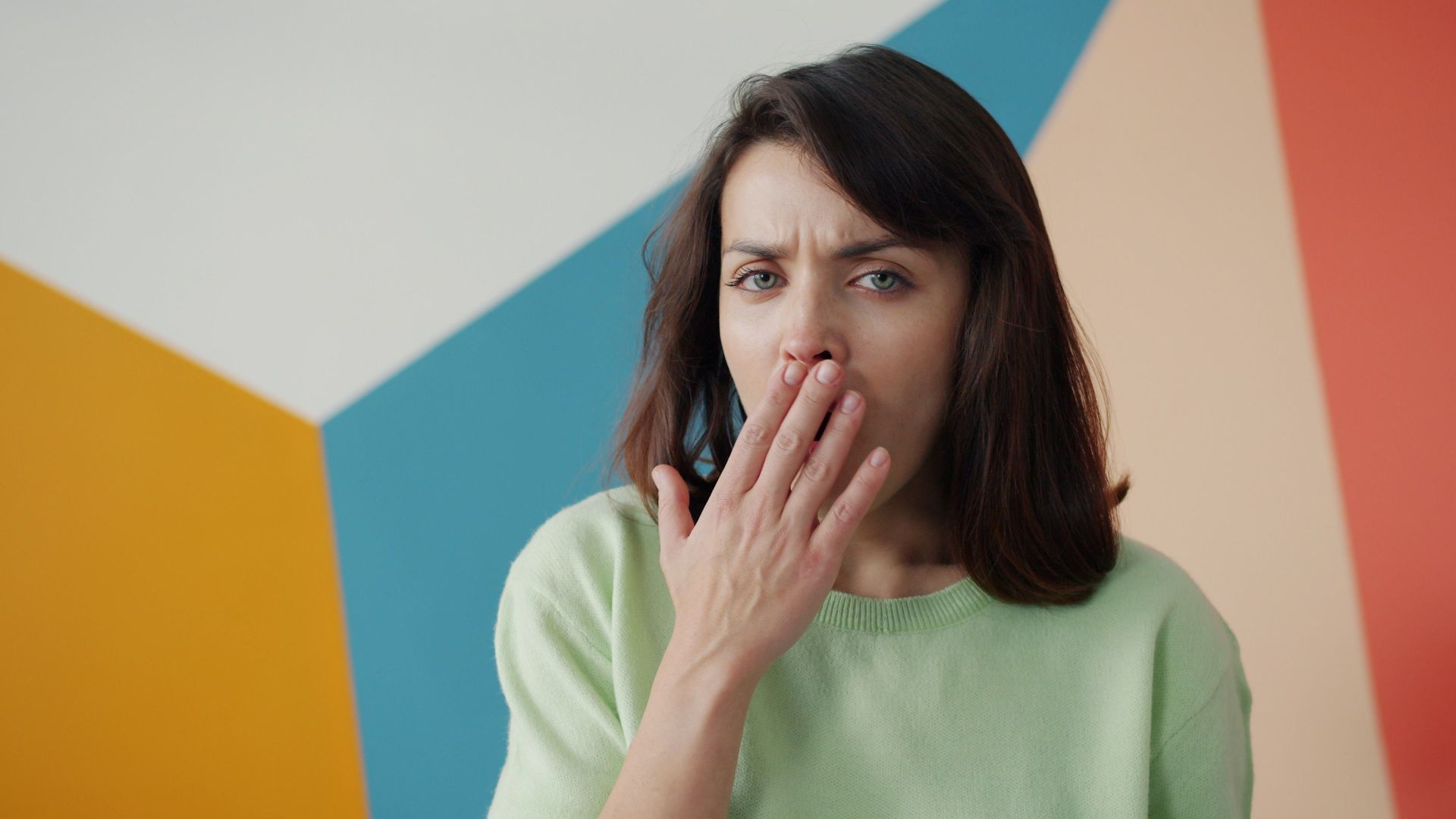 Woman covers her mouth against a colorful geometric wall, expressing surprise or yawn.