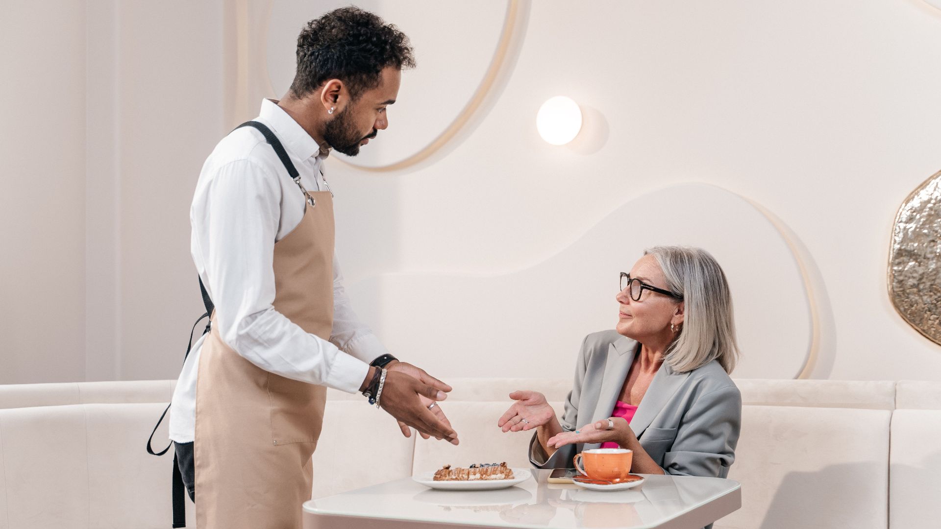 A waiter gestures while serving a senior woman in a stylish cafe setting, discussing dessert options.