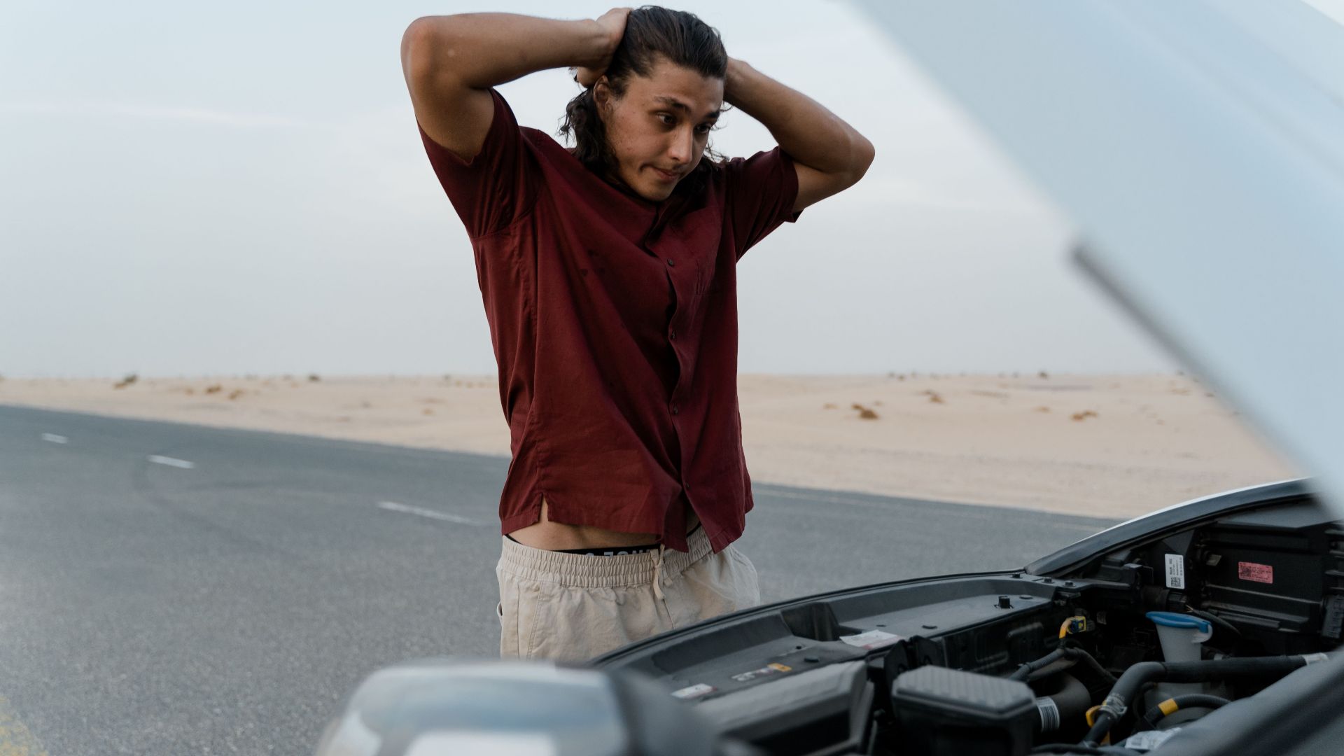 A frustrated man examining a broken car engine on a deserted road, showing stress and concern.