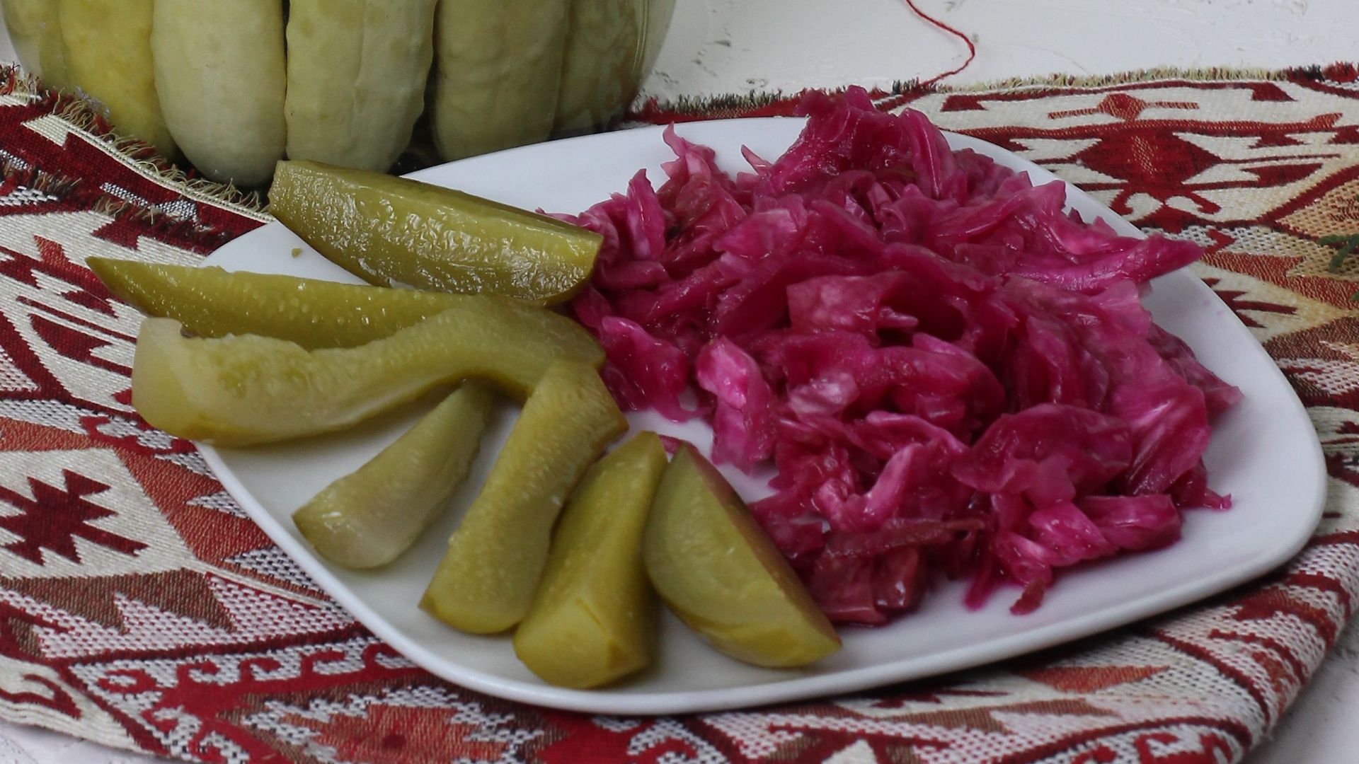 A jar of freshly pickled cucumbers with red cabbage plated on a patterned cloth, indoors setup.