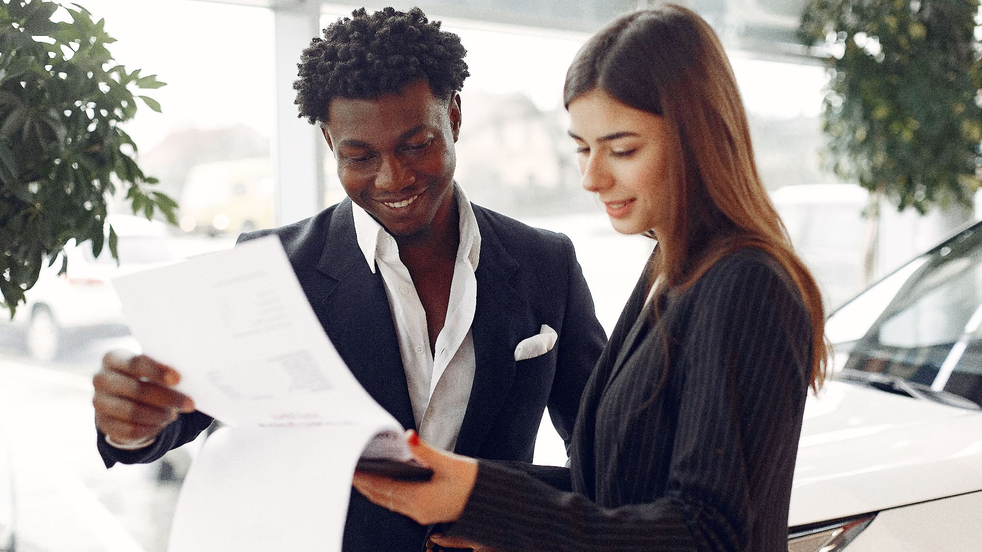 Cheerful smiling multiethnic businessman in classy suit and professional elegant female consultant standing close and reading contract details in car showroom