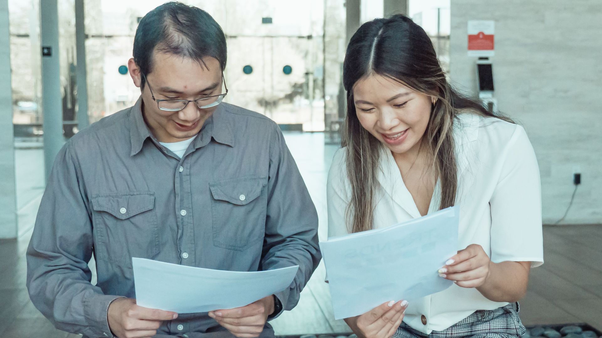 Two colleagues reading documents together on a bench in a modern office setting.