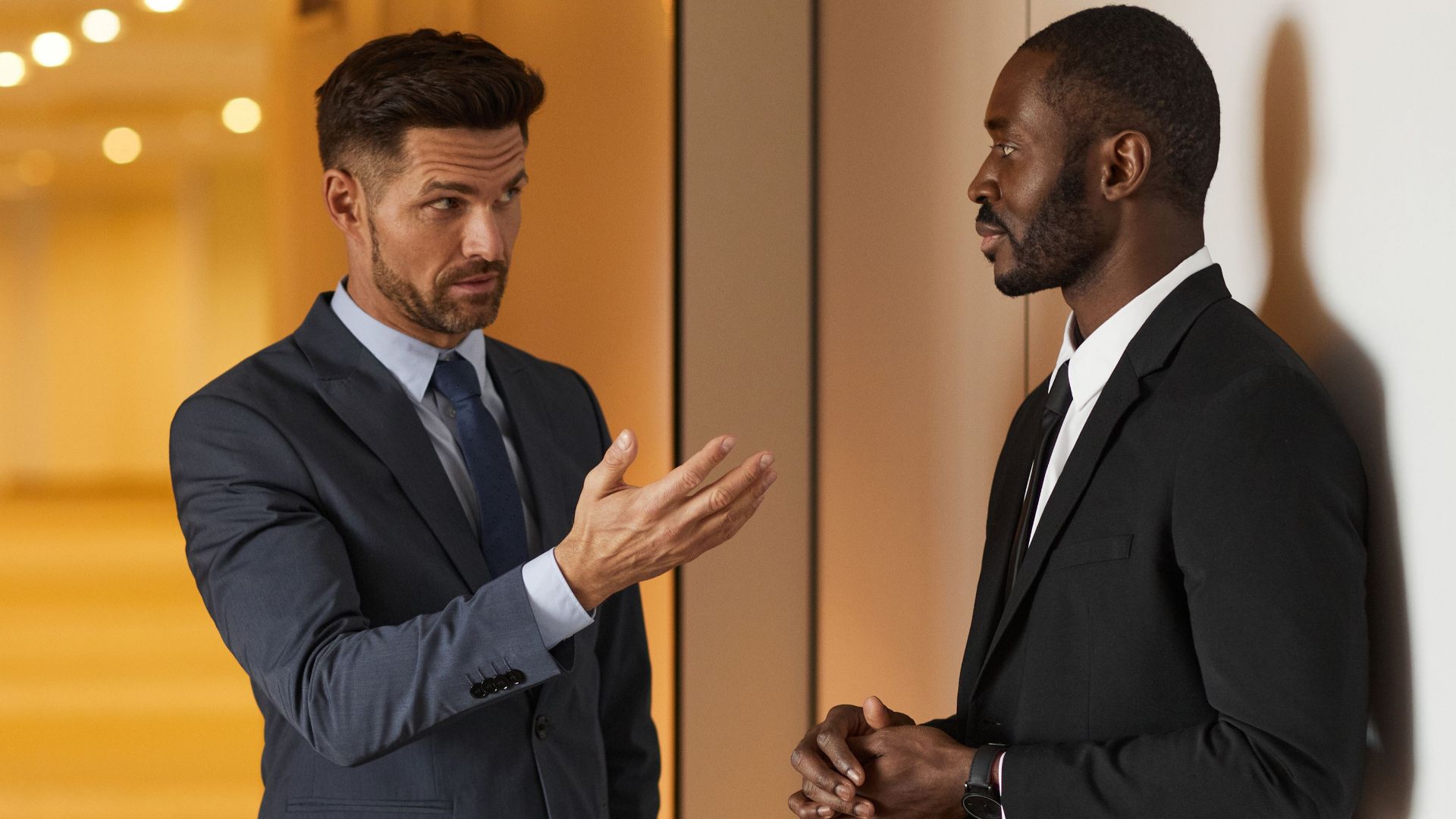 Two businessmen in formal attire engaged in a serious discussion in a modern office hallway.