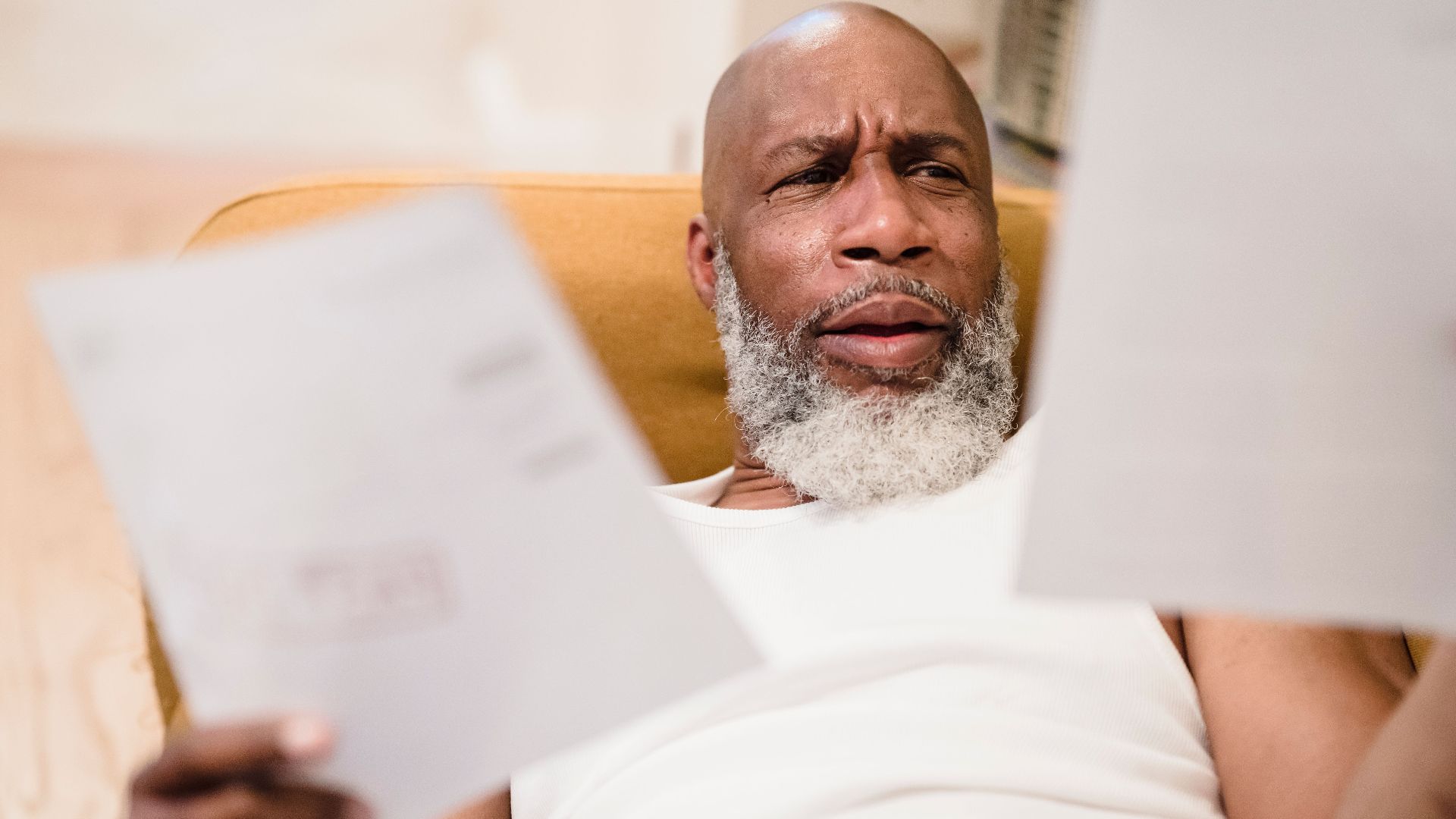 A middle-aged man looks concerned as he reviews household bills, sitting in a comfortable chair indoors.