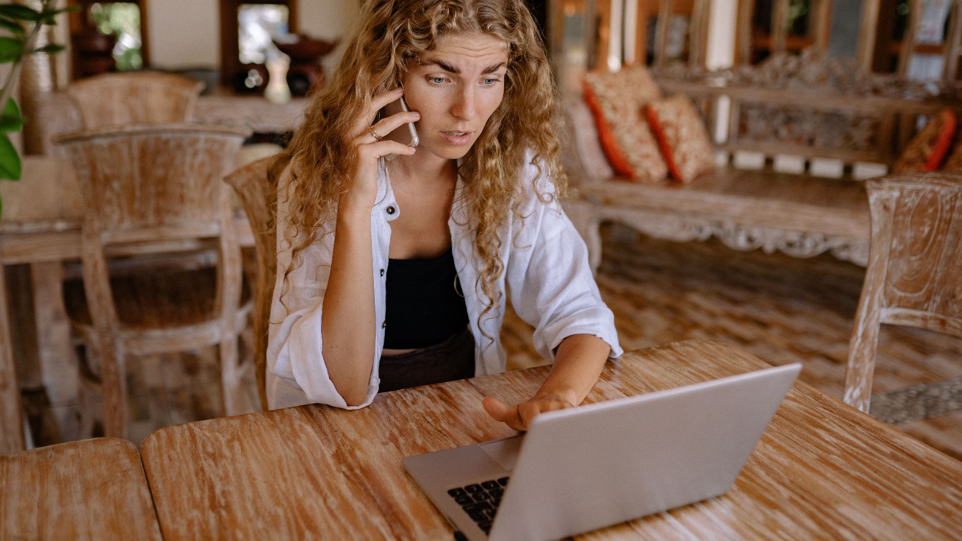 Woman sitting at wooden table, using laptop and talking on phone in a cozy indoor setting.