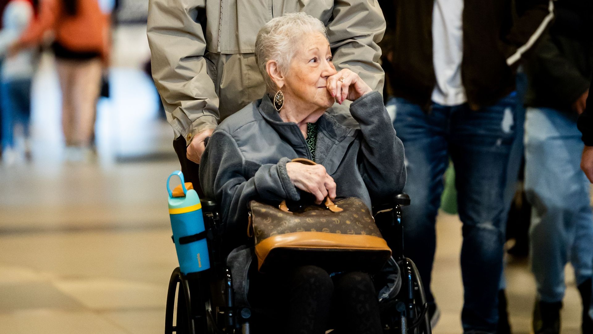 Senior woman in a wheelchair being assisted by a man in a bustling shopping mall.