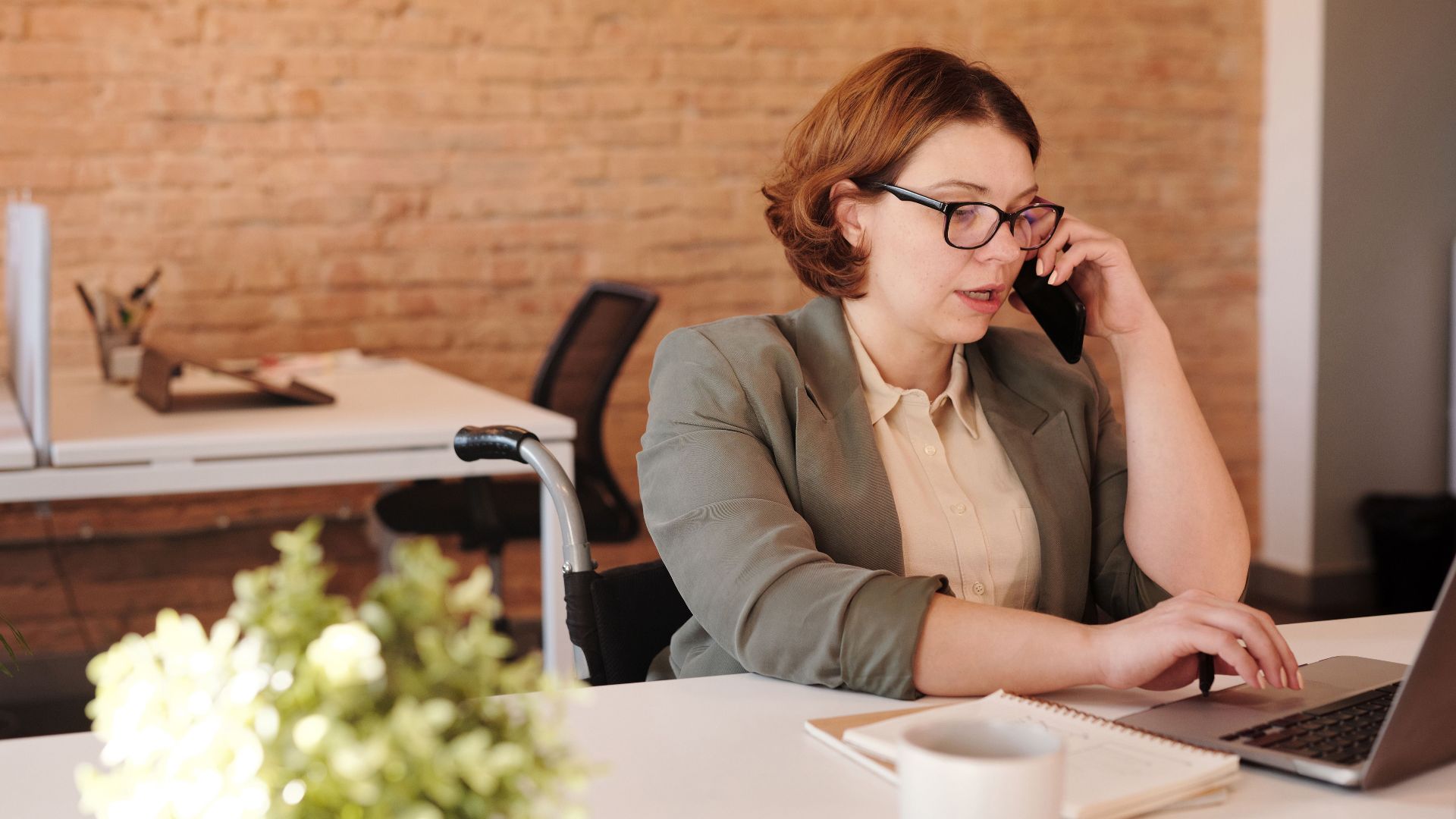 Professional woman in wheelchair working remotely on laptop and phone in a modern office.