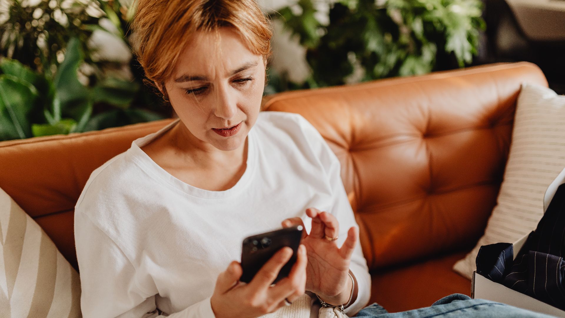 Woman in casual attire using a smartphone on a cozy sofa surrounded by plants.