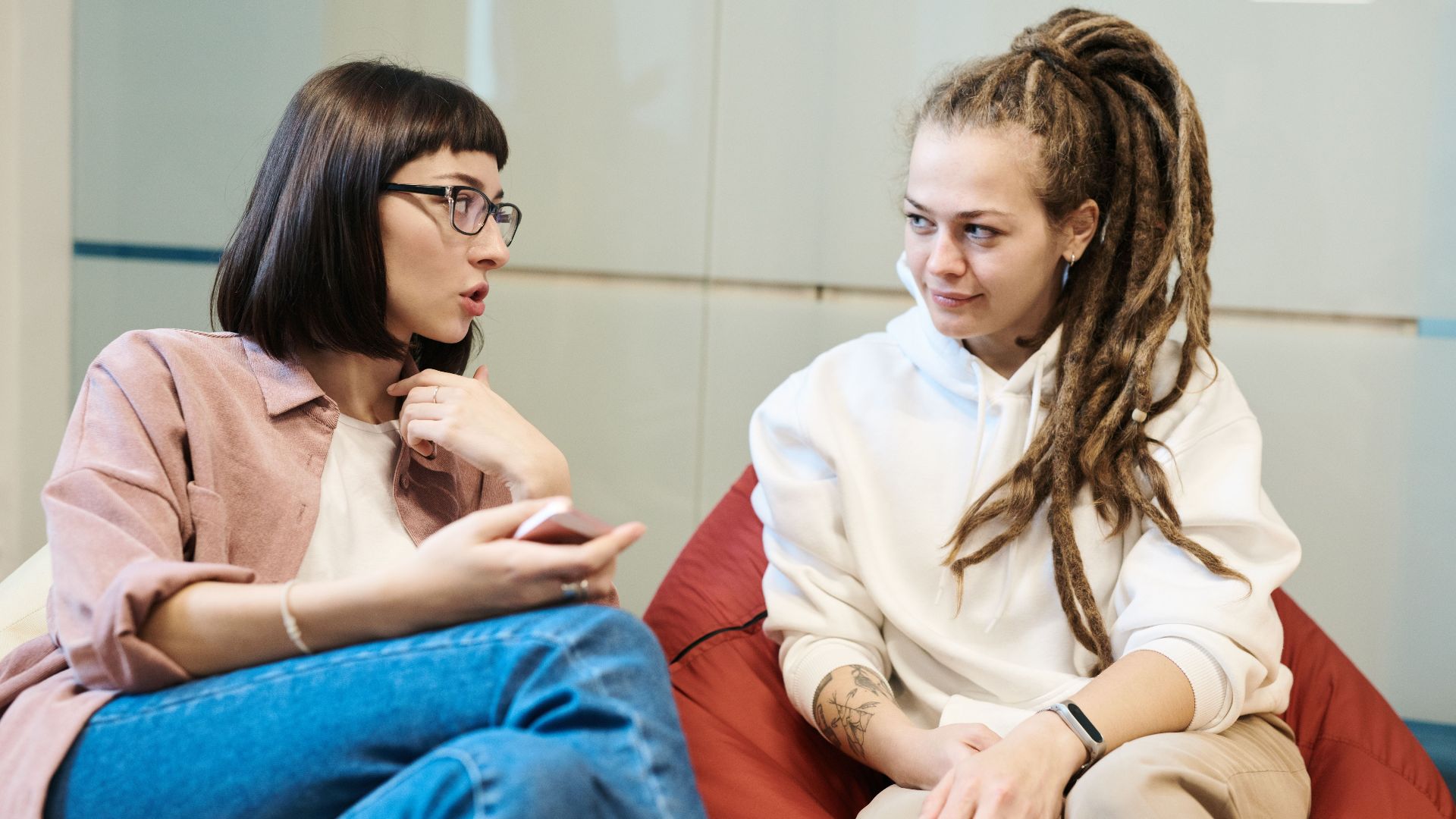 Two women engaged in a conversation in a modern office, sitting comfortably in relaxed chairs.