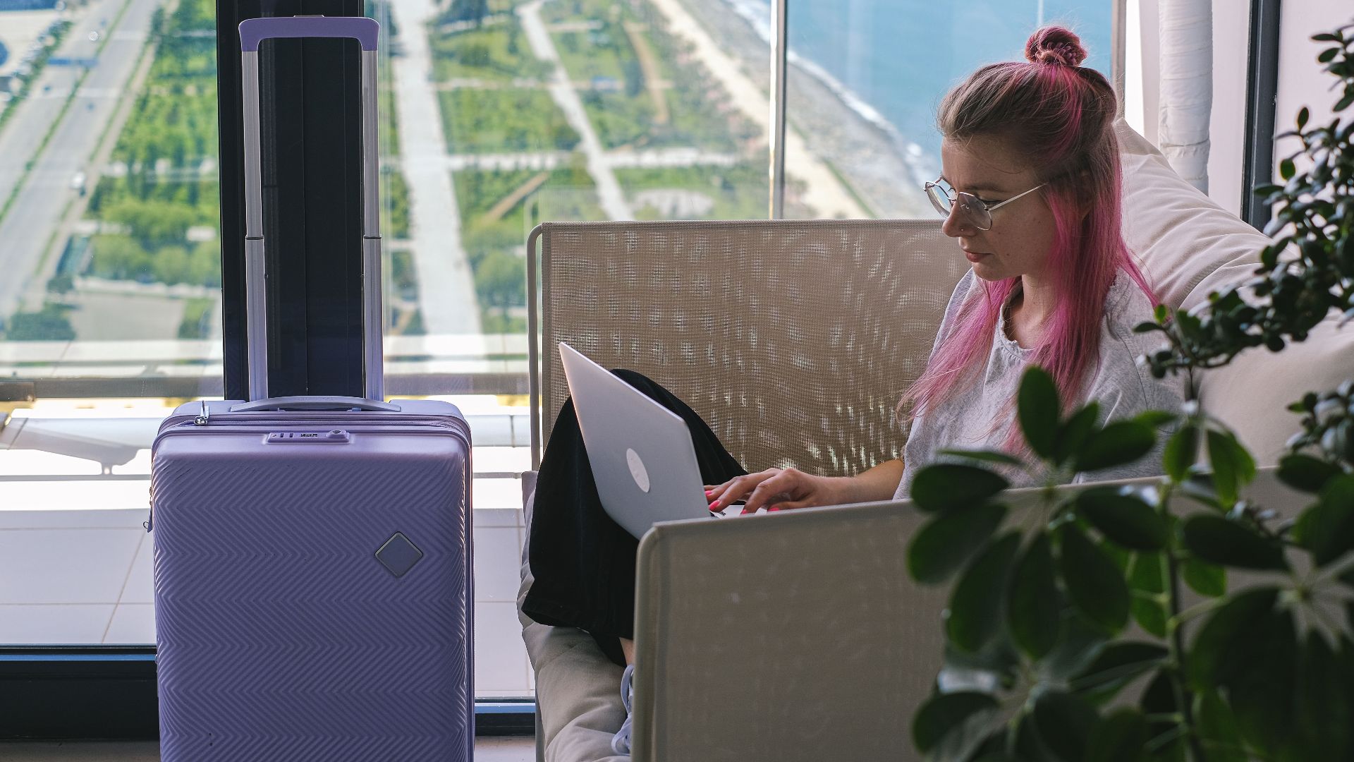 Woman with pink hair working on laptop in hotel room overlooking scenic landscape. Travel and freelance concept.