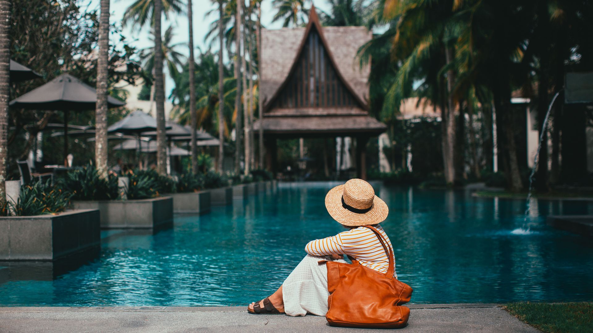 Side view anonymous female wearing summer clothes and hat resting on poolside in tropical resort amidst lush palms and bungalows