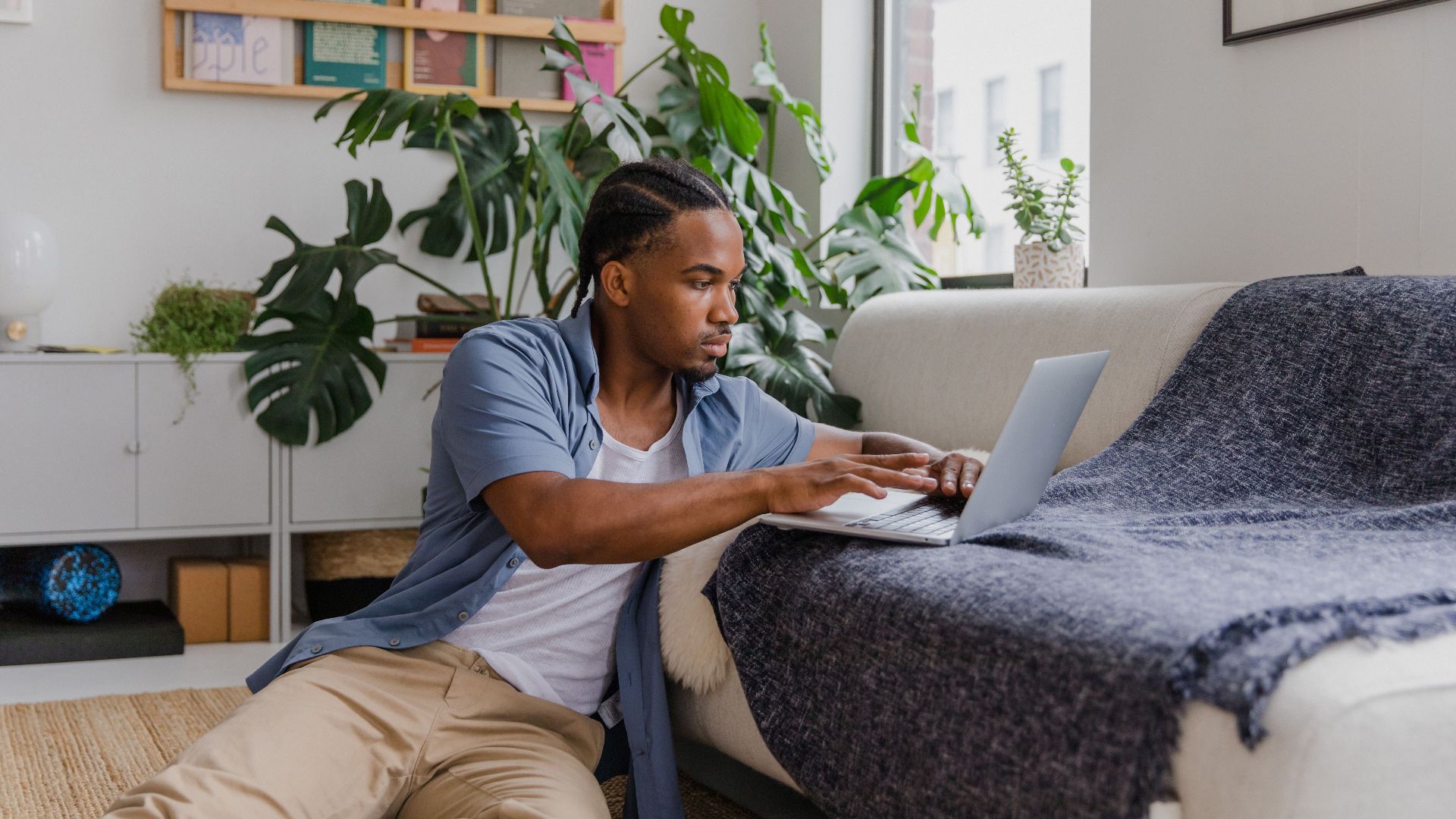 Man working on laptop while sitting on floor in a stylish, plant-filled living room.