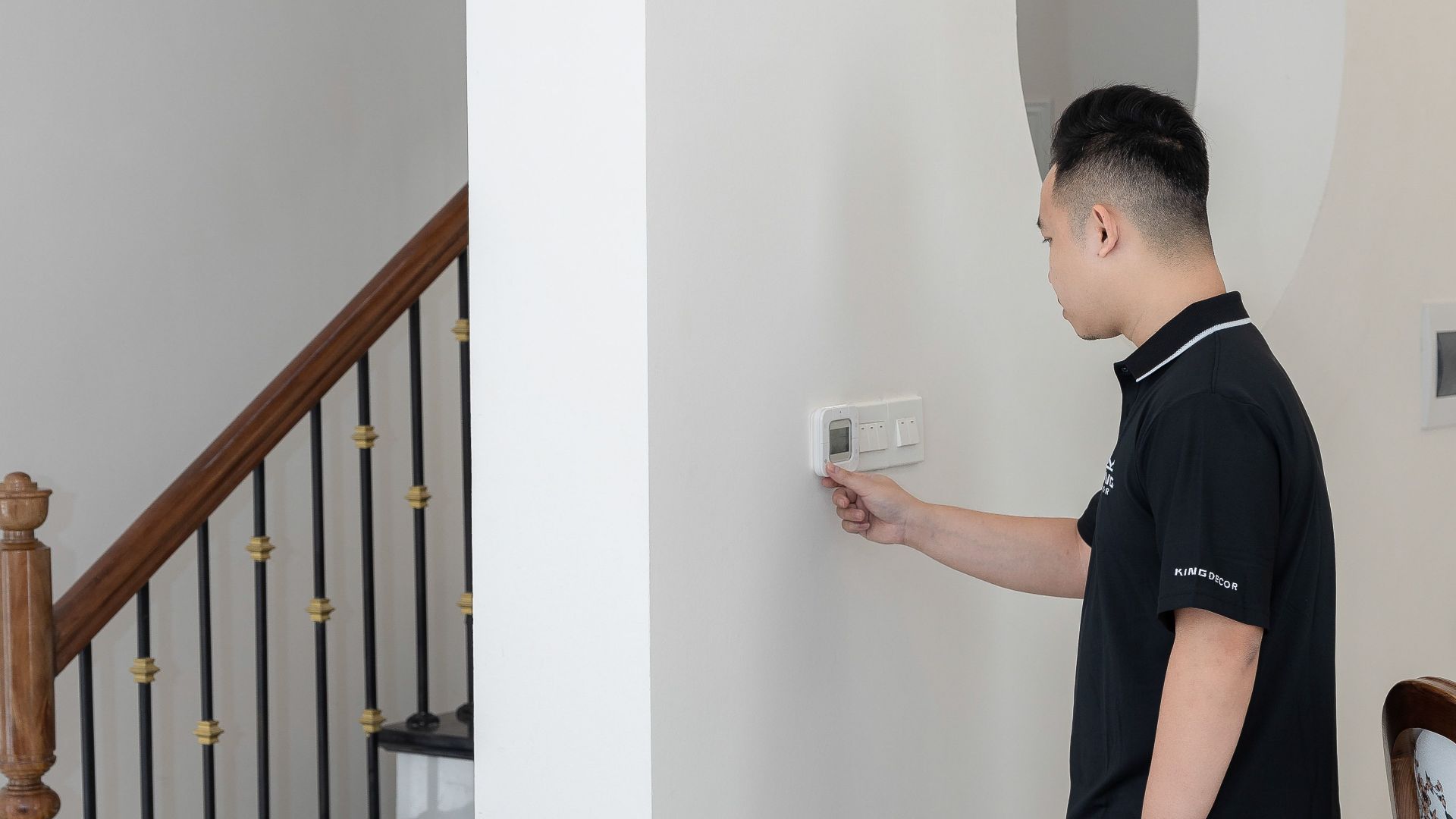A young man adjusts the thermostat on a wall in a modern indoor setting with a staircase.