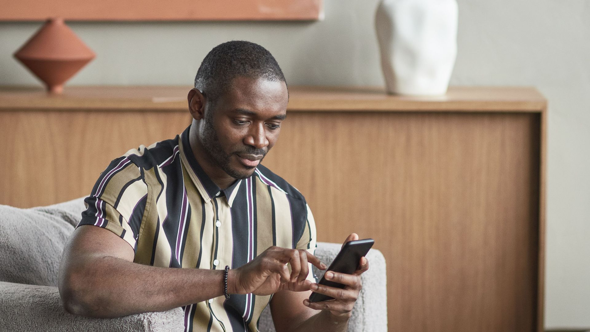 Casually dressed man sitting in armchair using smartphone in modern living room.