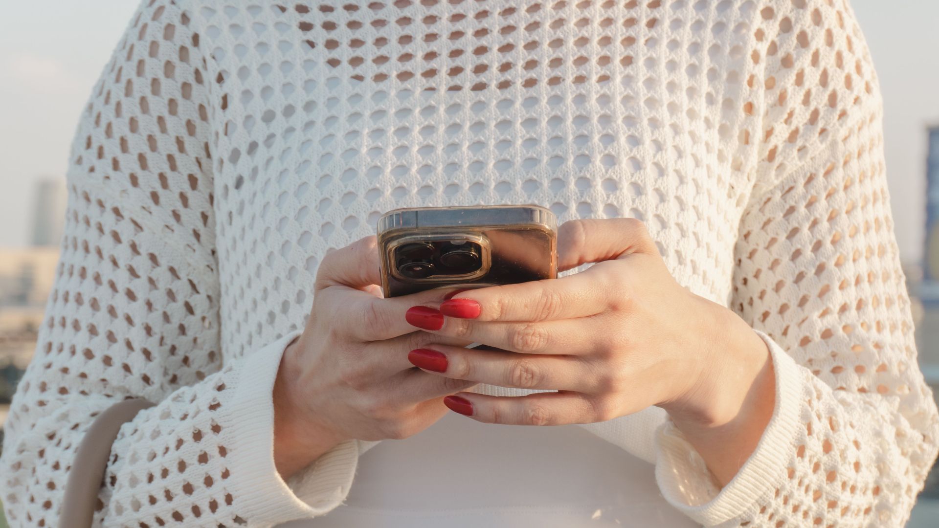 Close-up of a woman using a smartphone outdoors, wearing a stylish casual outfit with red nails.