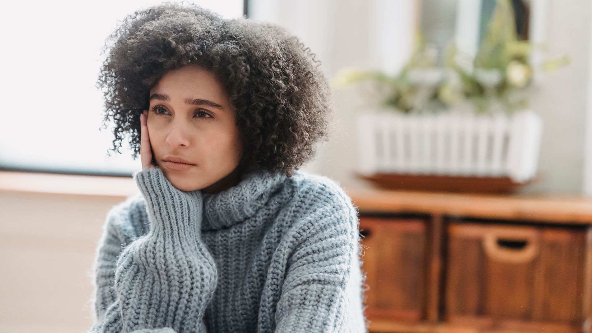 Discontent African American female leaning on hand and looking away while sitting in light room near green plant on blurred background