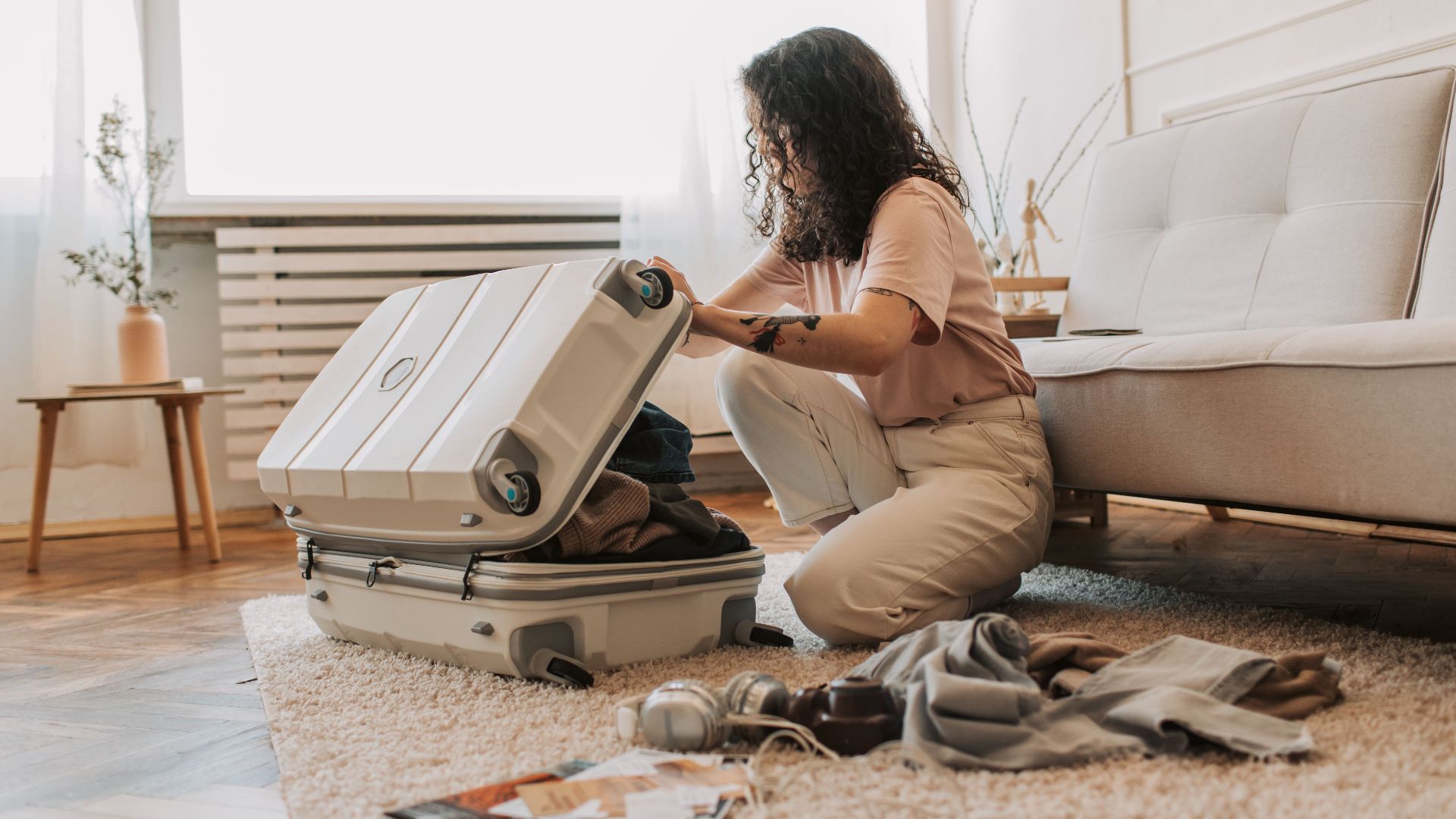 A woman packing her suitcase in a stylish living room, surrounded by clothes and travel accessories.