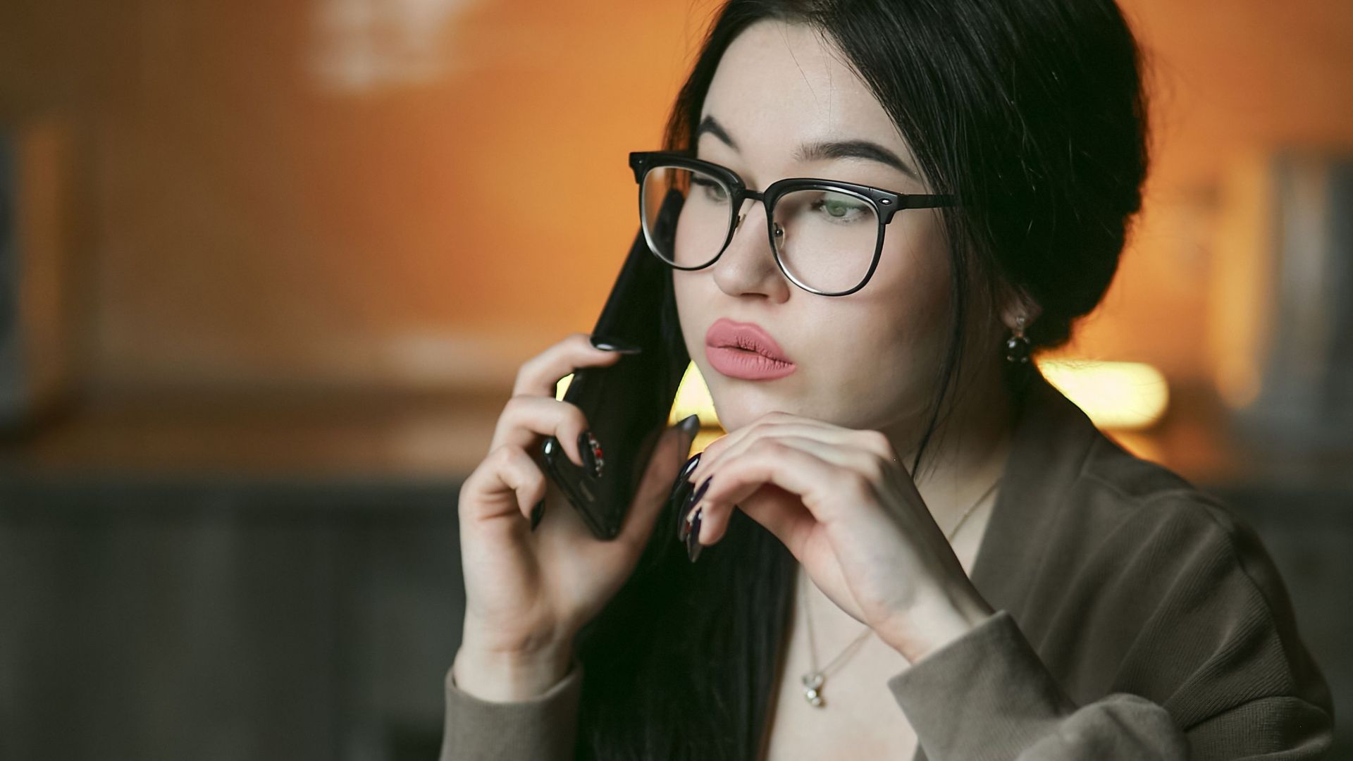 A woman with eyeglasses engaged in a phone call indoors, focused and thoughtful.